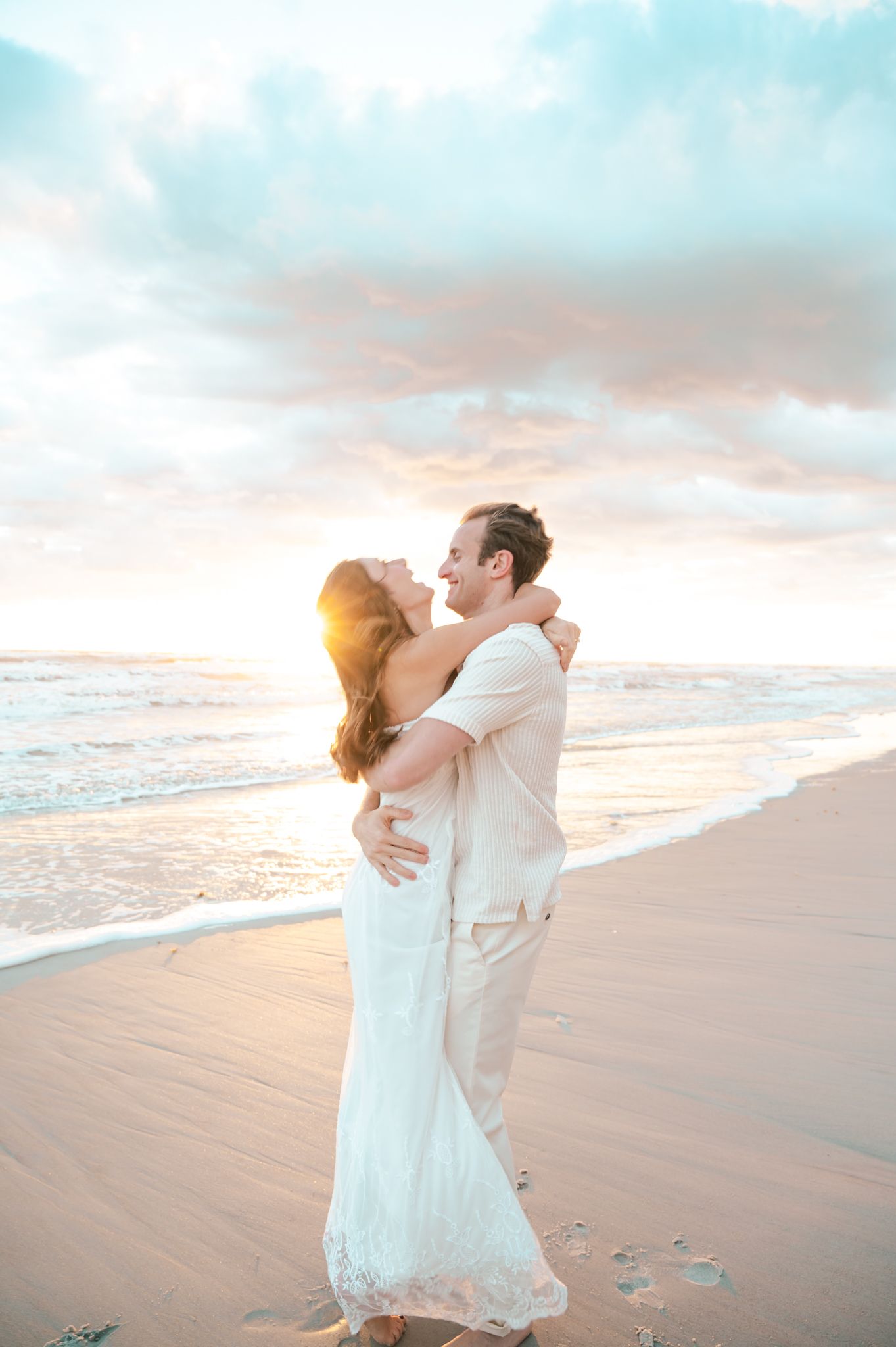 Couple embracing on a sandy beach at sunset, woman in white lace dress, golden light reflecting on ocean waves.