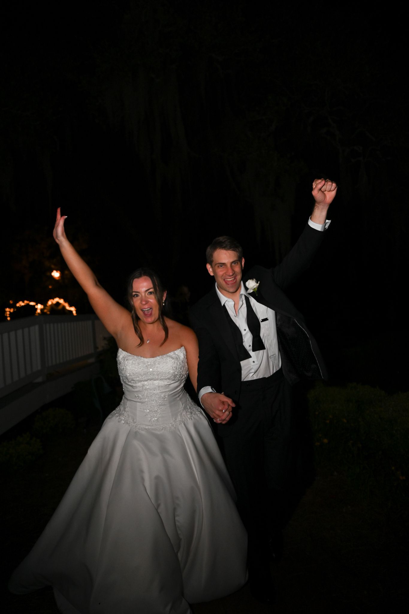 Joyful bride and groom celebrate outdoors at night, raising fists in triumph wearing wedding attire.