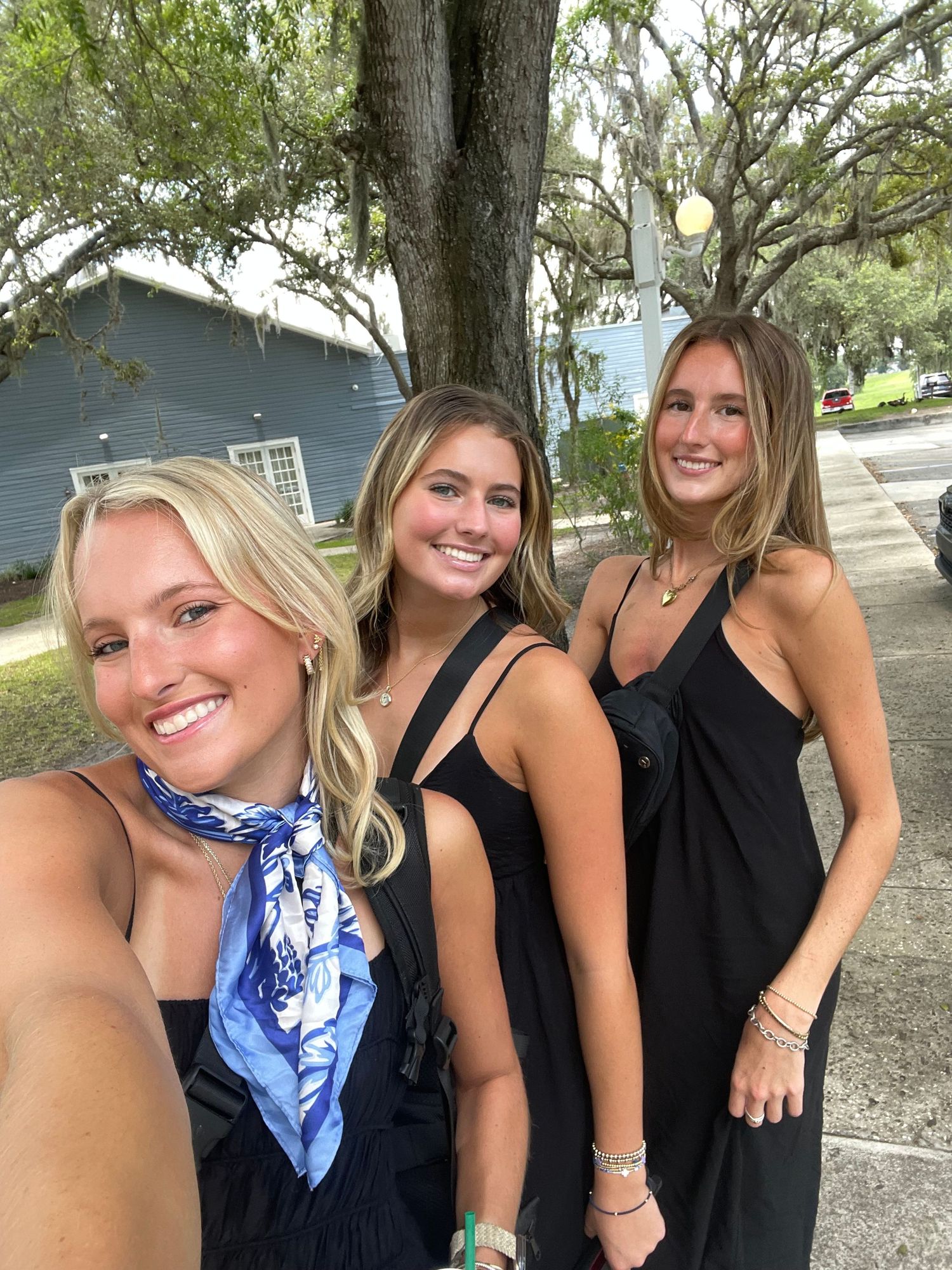 Three young women in black outfits smile for a selfie outdoors near a tree-lined street.