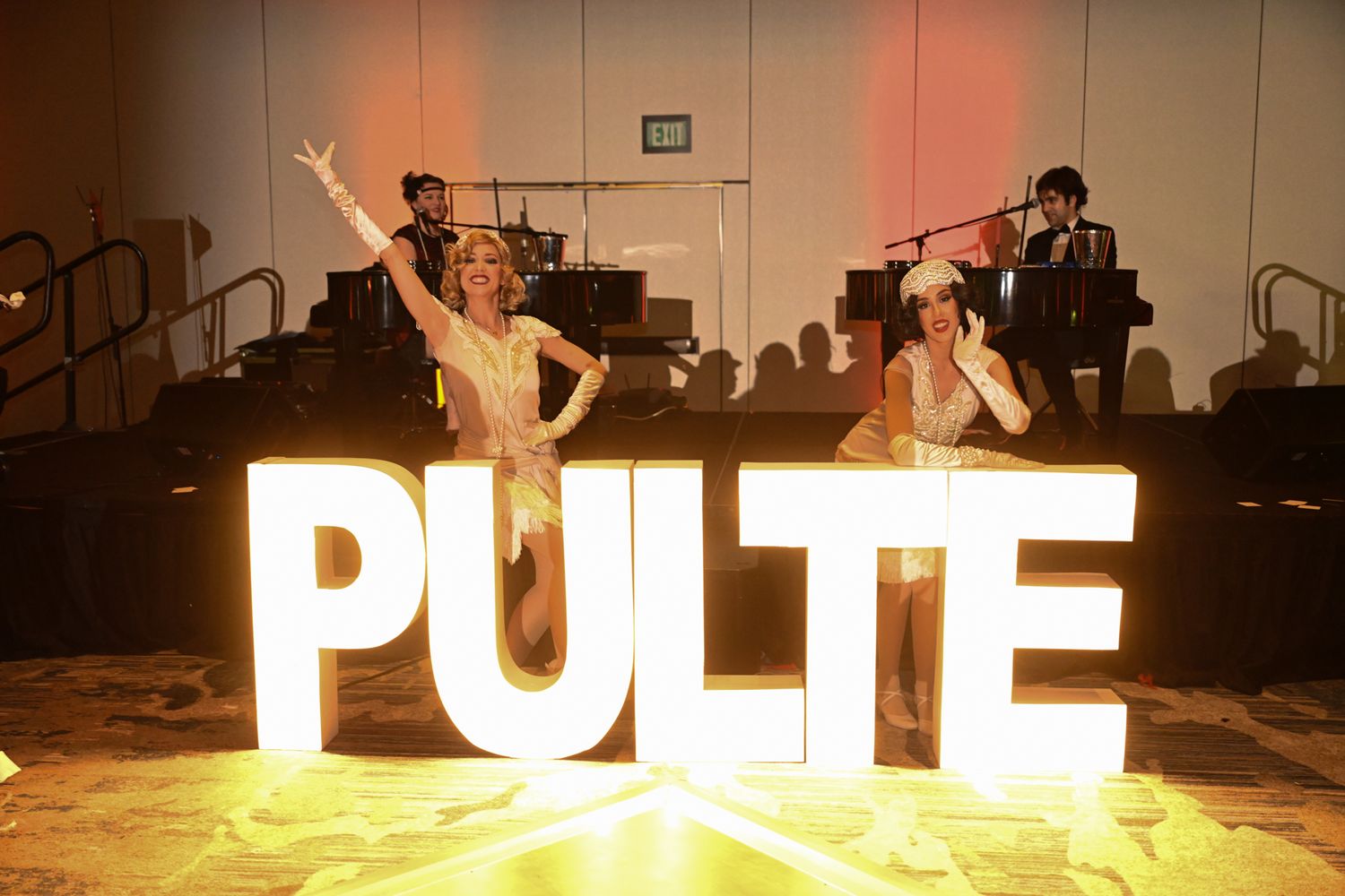 Two performers dance beside illuminated PULTE marquee letters at a corporate gala event with a live band.