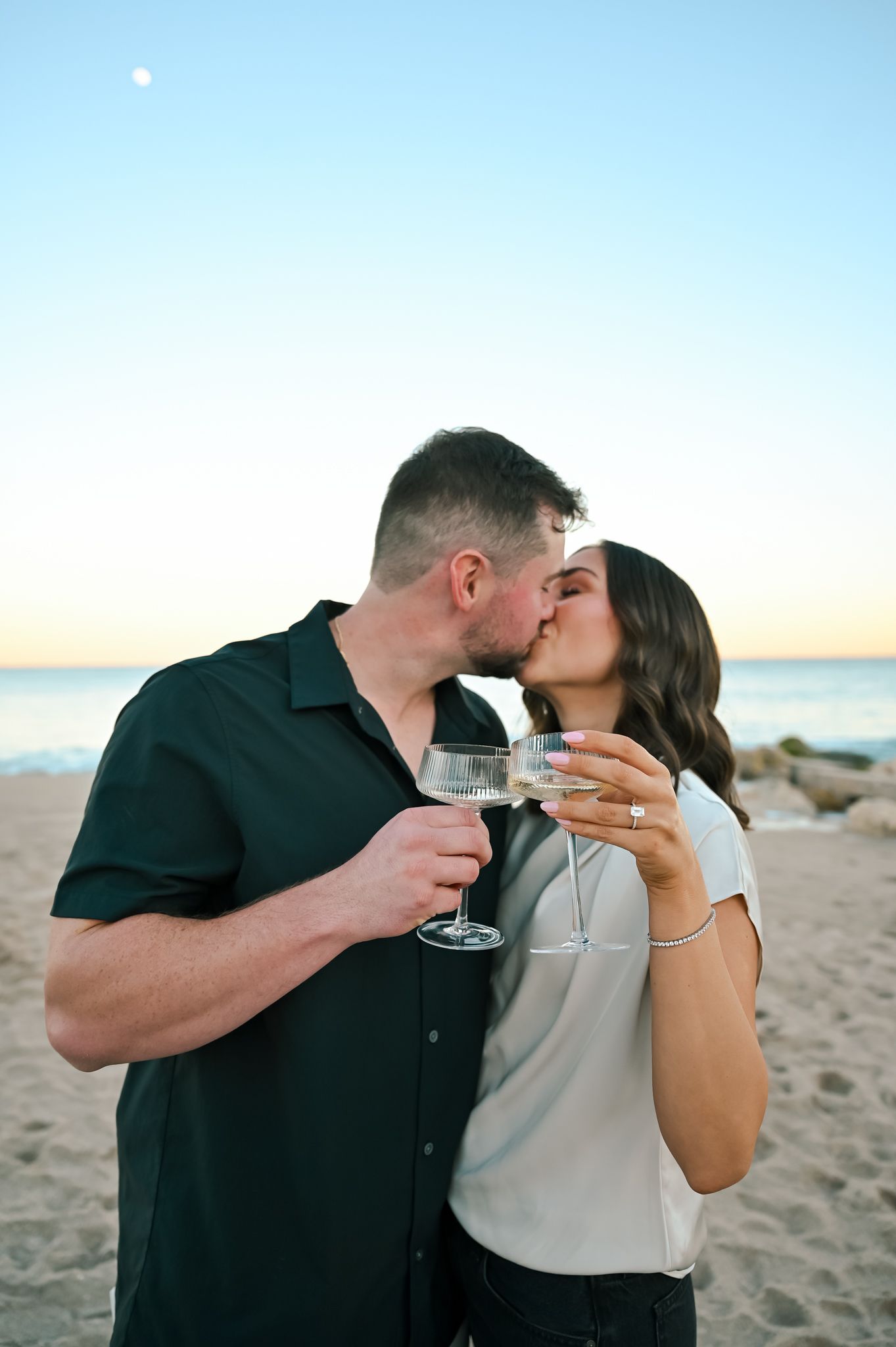 Couple kissing on beach at sunset while holding champagne glasses, celebrating engagement with ocean backdrop.
