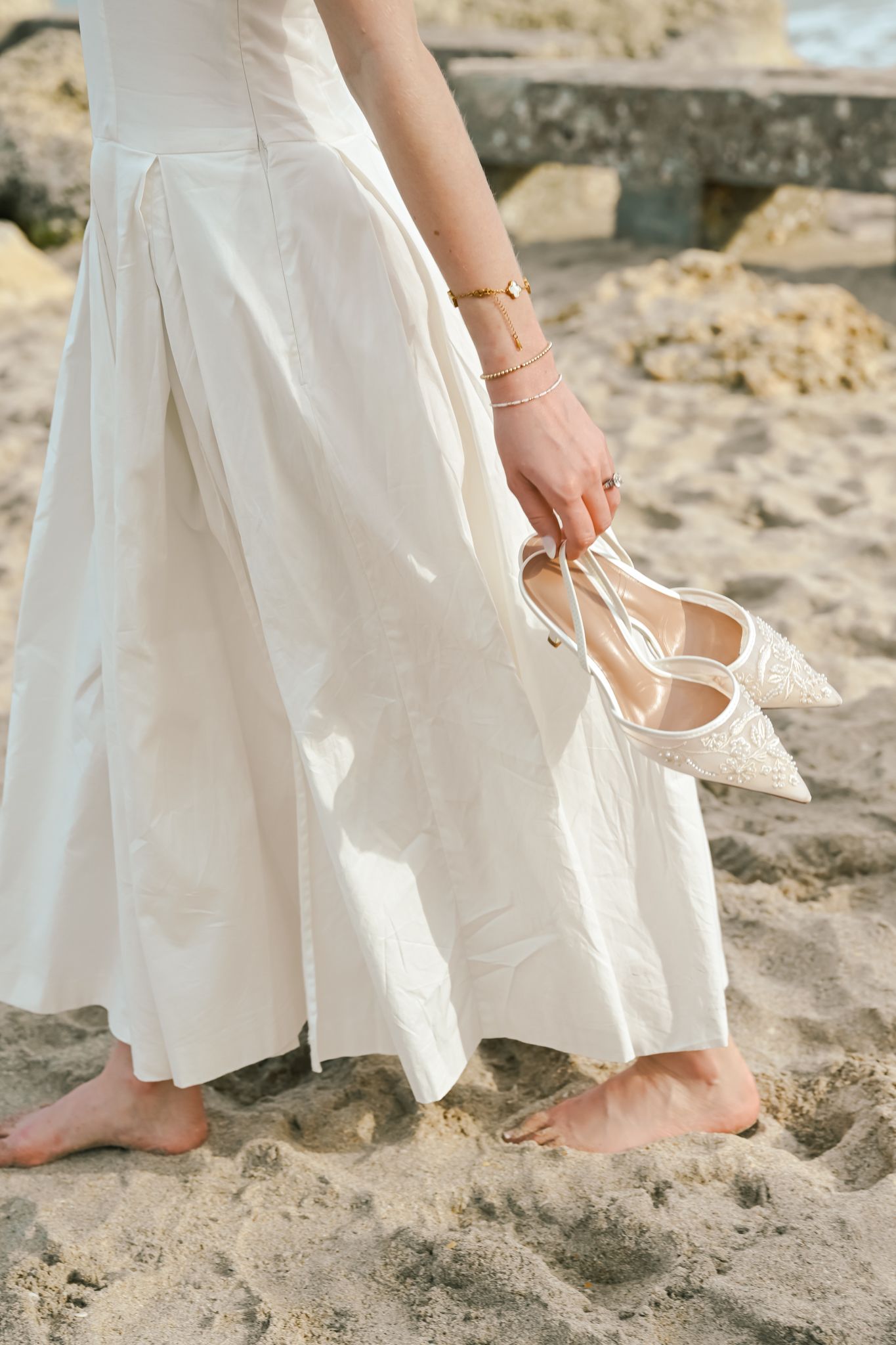 Barefoot bride in white dress holding pearl wedding heels on sandy beach.