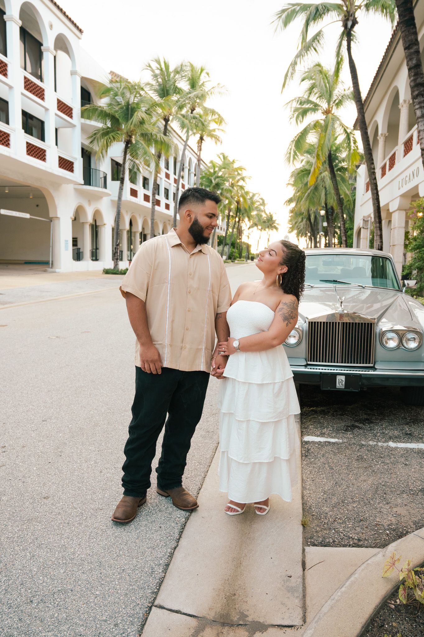 Couple in white dress and beige shirt holding hands beside a classic Rolls-Royce, palm trees and resort building behind them.