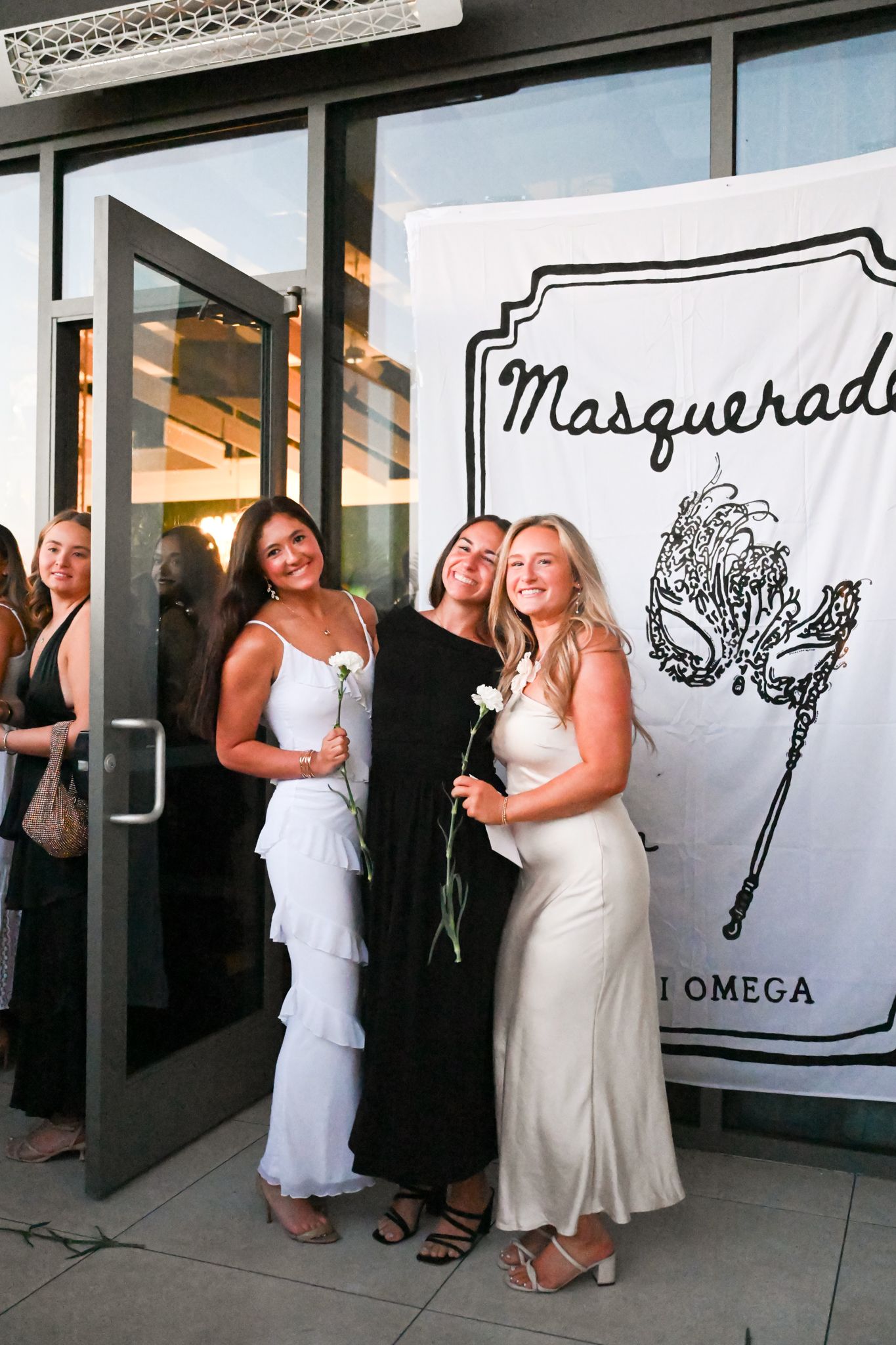 Three women in elegant dresses pose in front of a Chi Omega Masquerade event banner, holding white roses.
