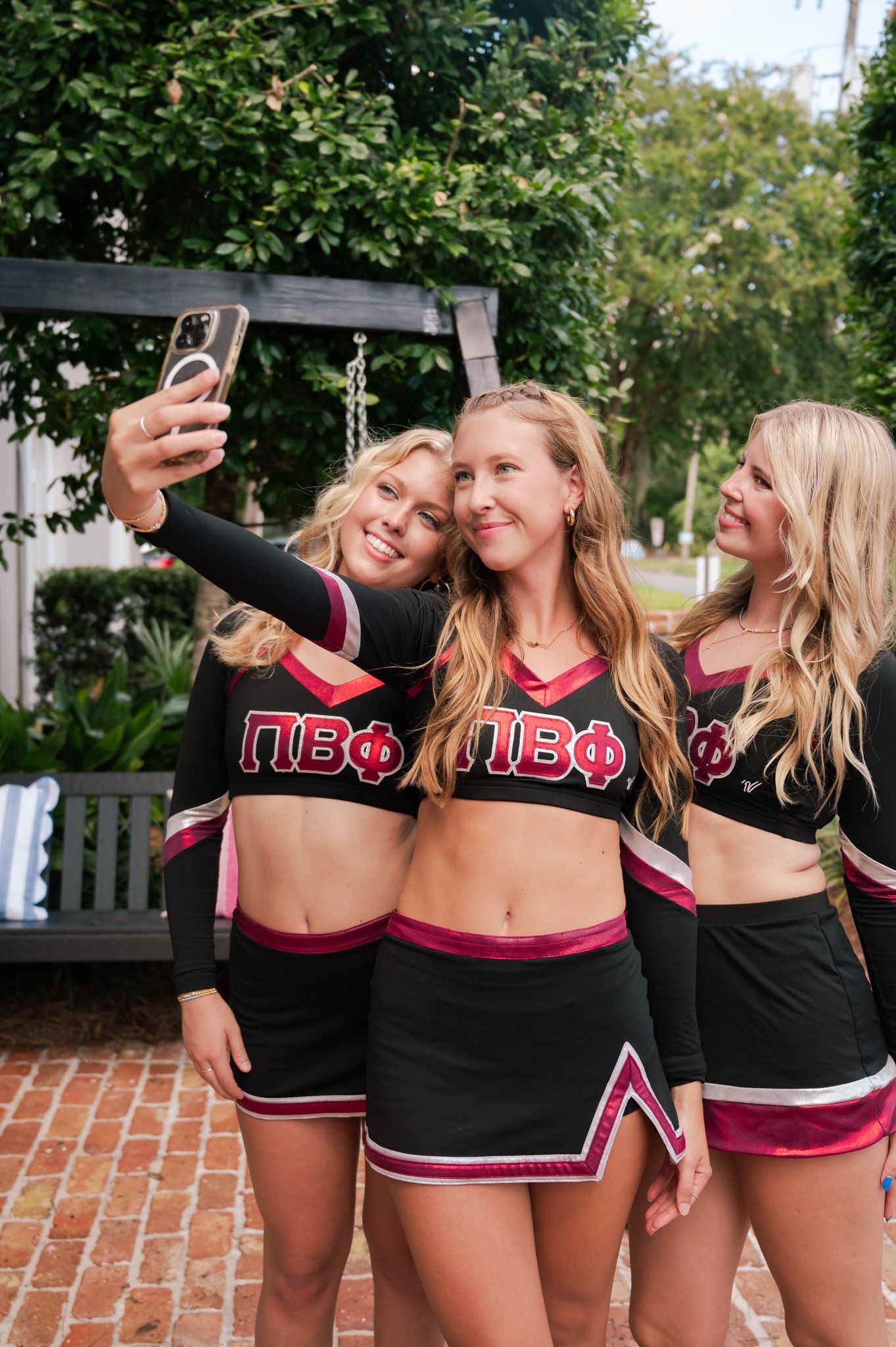 Three blonde women in Pi Beta Phi cheerleader uniforms taking a selfie outdoors.