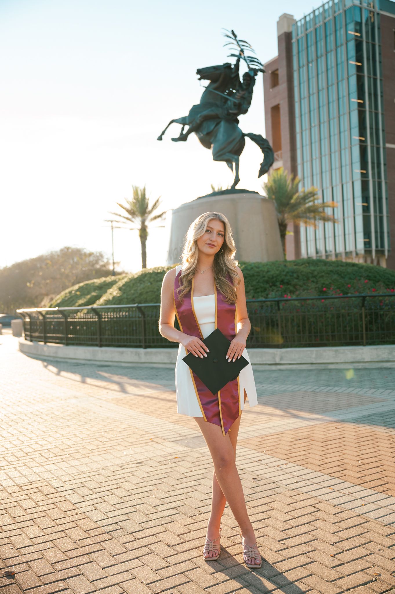 Graduate poses with university statue in background during sunset photo session.