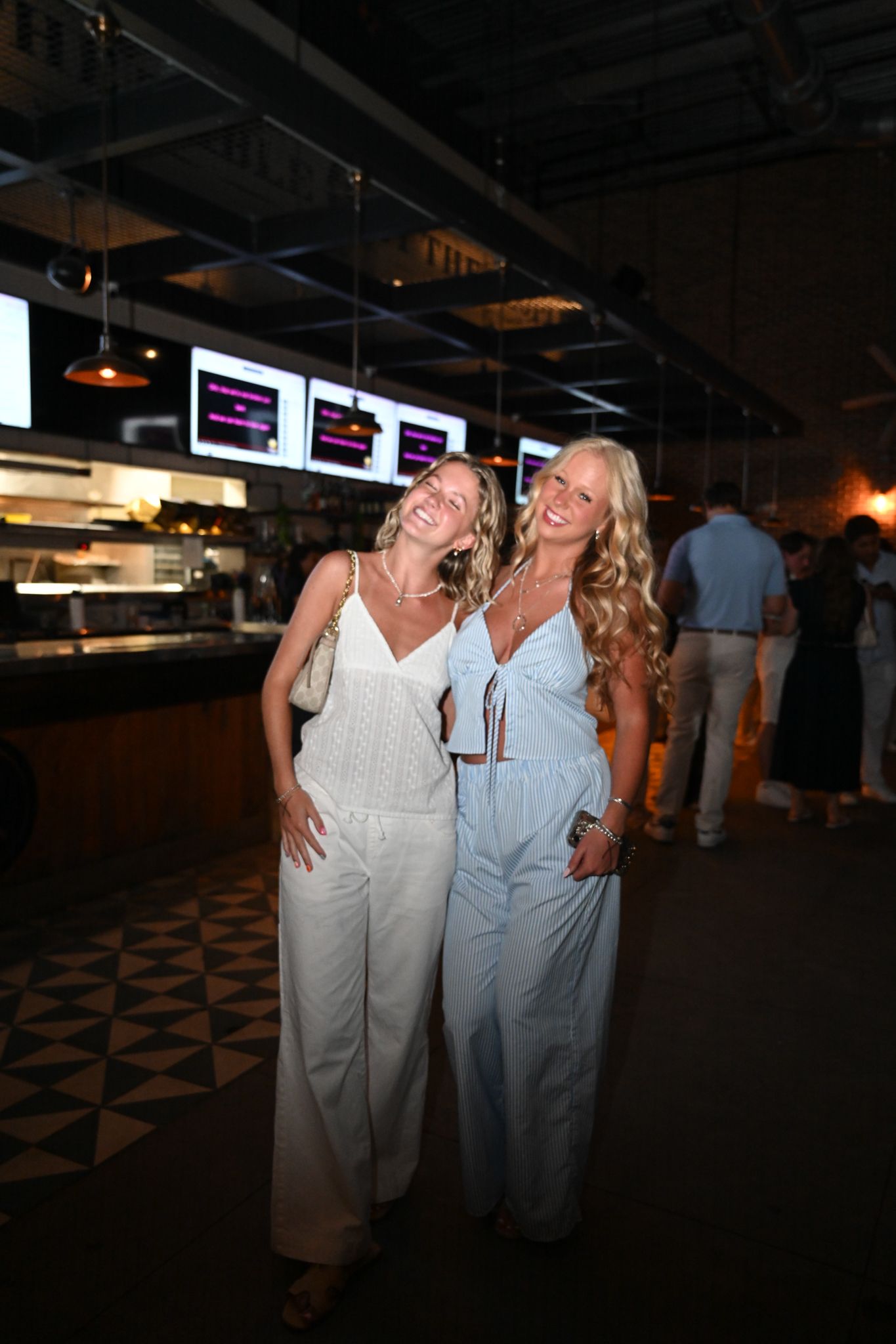 Two smiling women in white and light blue jumpsuits pose together inside a modern restaurant bar.