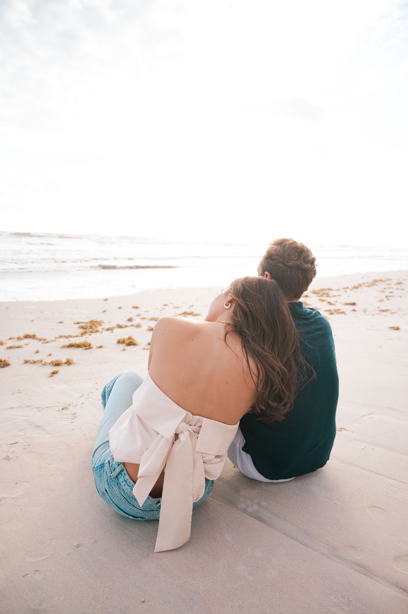 Couple sitting together on a sandy beach, gazing at the ocean, woman in off-shoulder top and jeans.