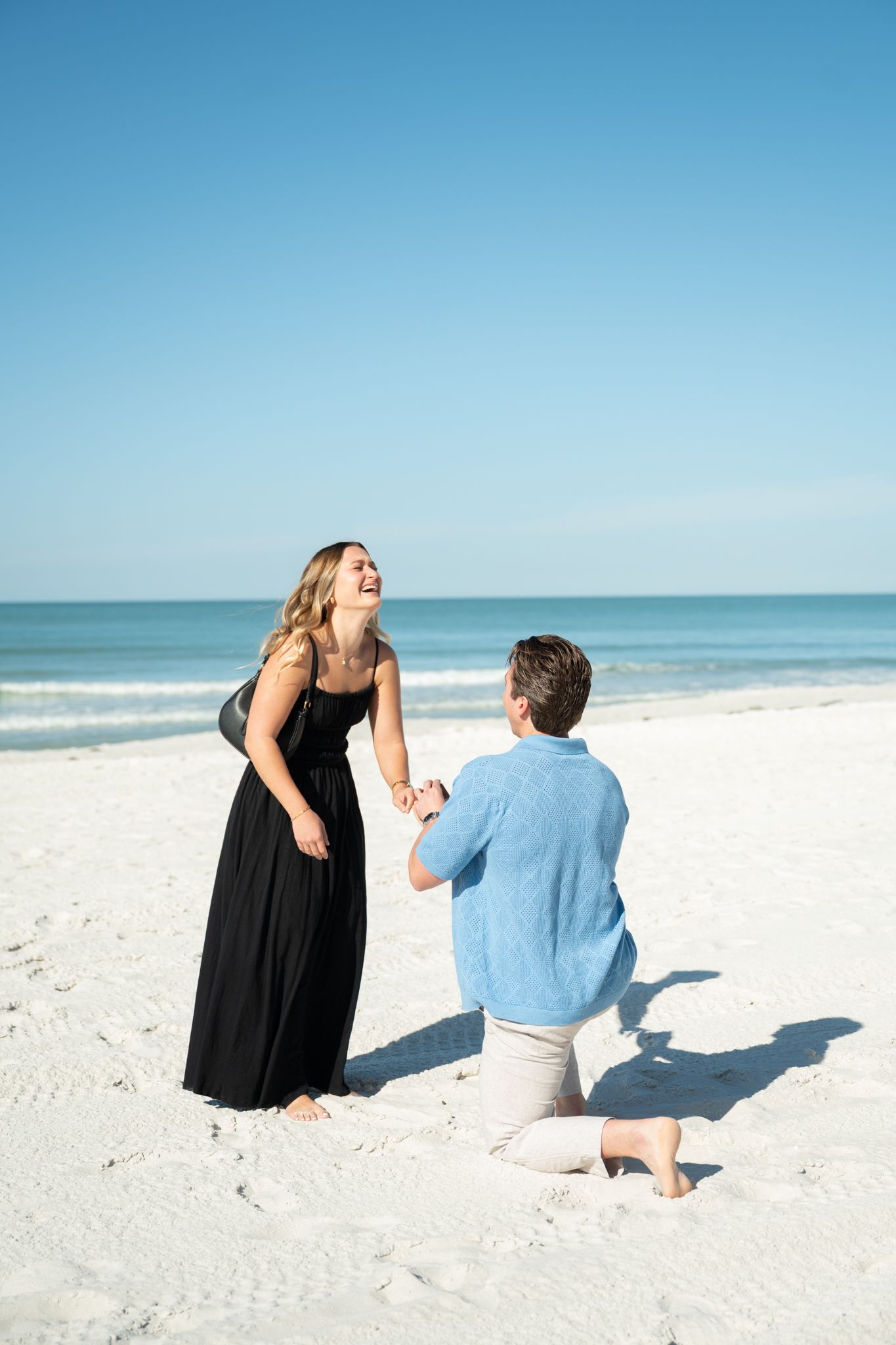 Man proposing on one knee to woman in black dress on a sunny white sand beach with ocean backdrop.