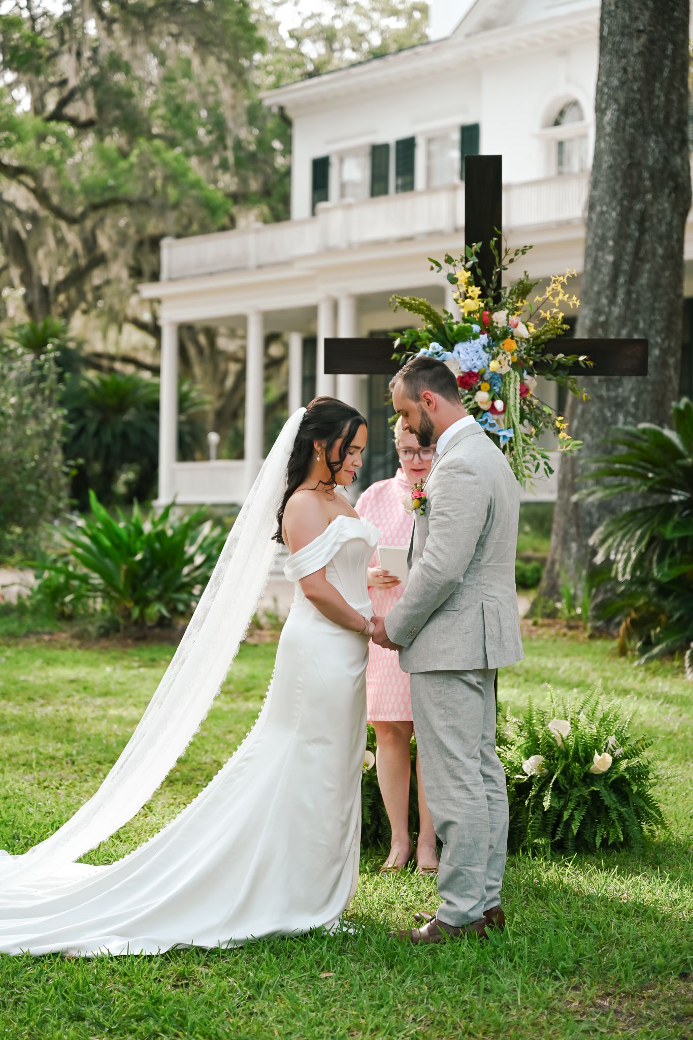 Bride and groom exchange vows outdoors before a floral cross at an elegant Southern estate wedding ceremony.