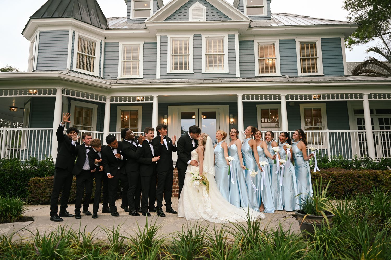 Wedding party poses in front of a blue Victorian mansion, bridesmaids in light blue gowns, groomsmen in black tuxedos.