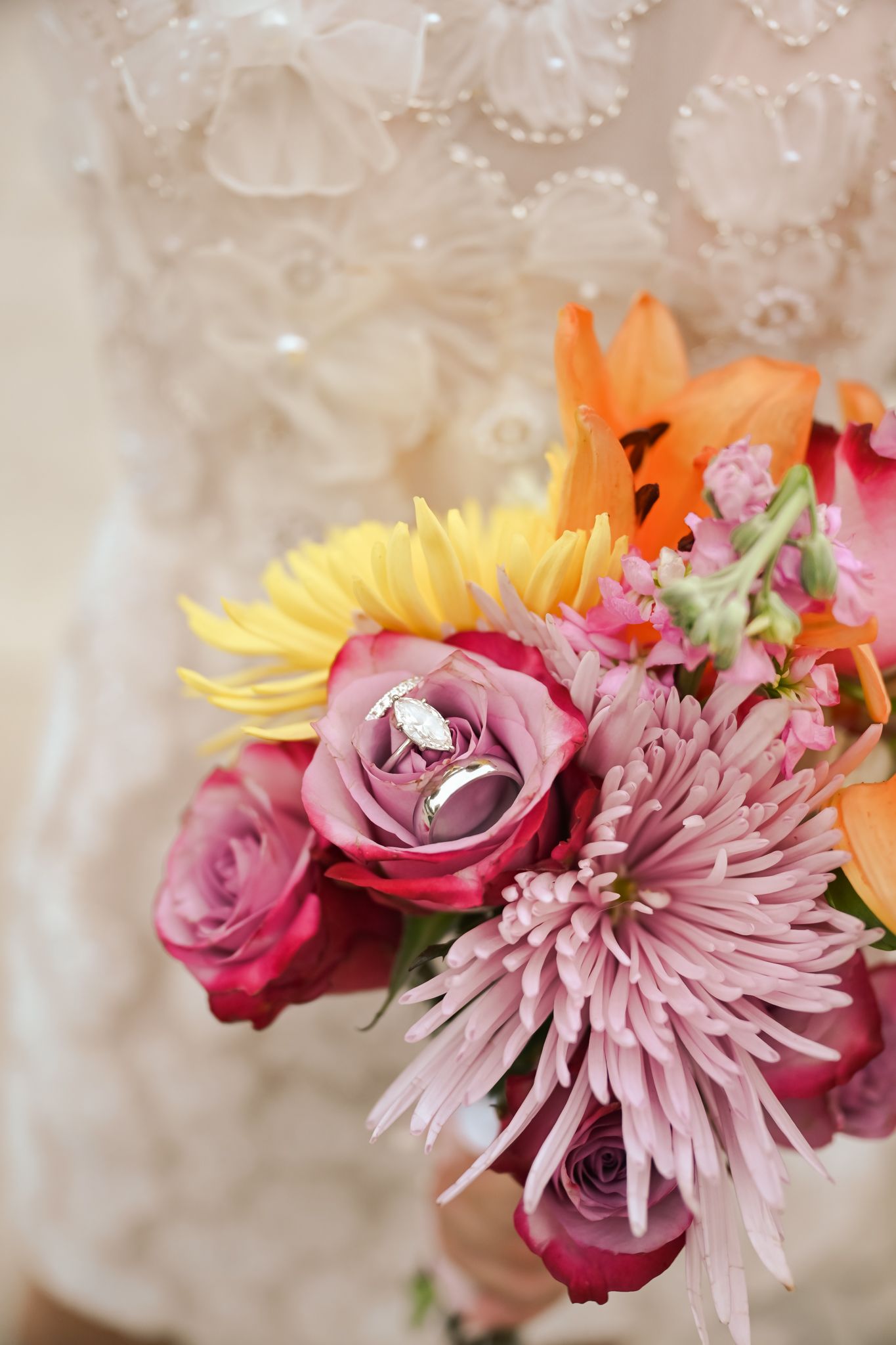 Wedding rings rest on a colorful bridal bouquet with pink roses, chrysanthemums, yellow sunflowers, and orange lilies.