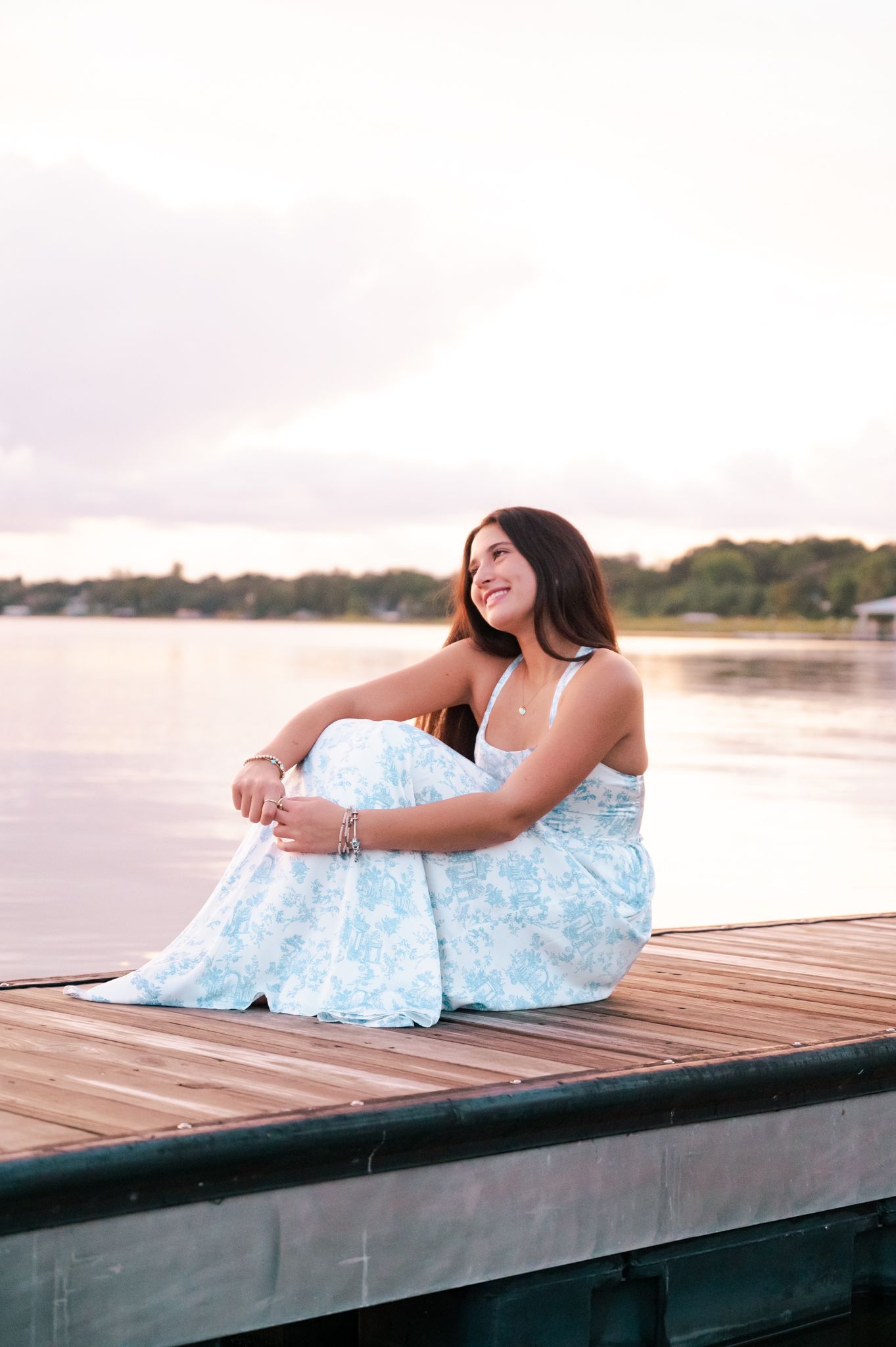 High School senior in light blue dress relaxes on wooden dock by peaceful waterfront.