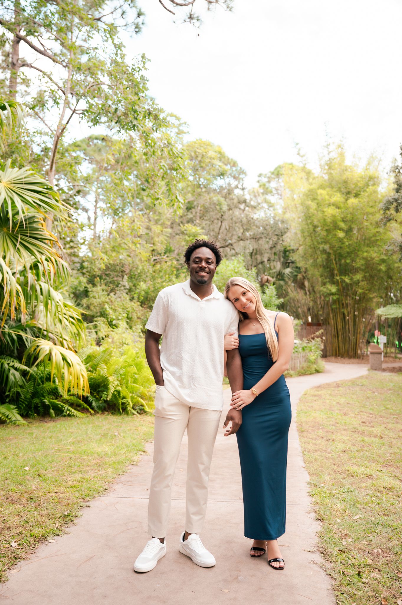 A couple poses on a garden path, man in white outfit and woman in teal dress, surrounded by lush tropical greenery.