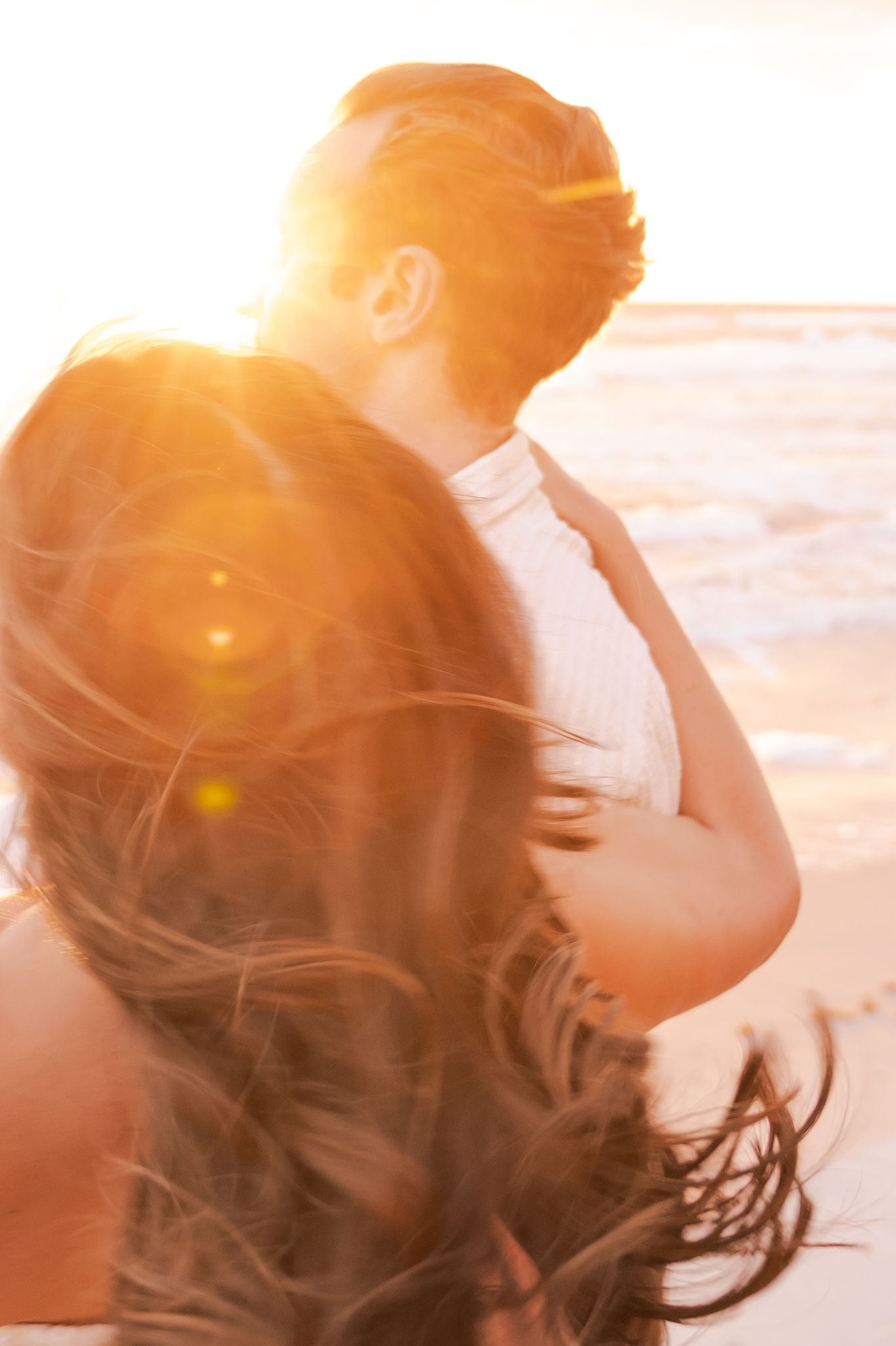 Couple embracing on beach at golden sunset with warm lens flare and flowing hair in romantic outdoor portrait.