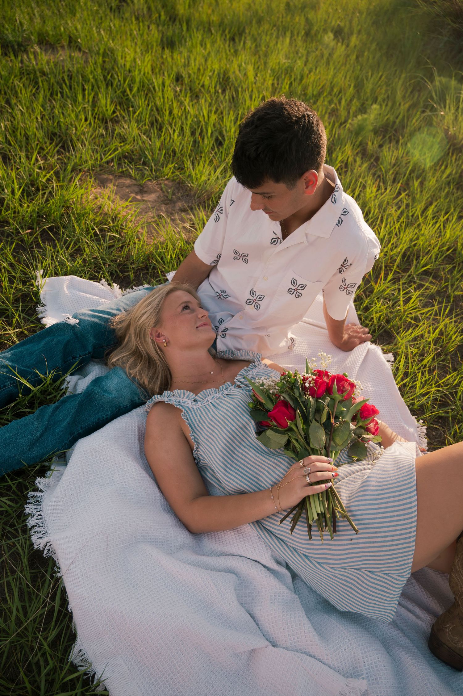 Romantic couple relaxing in green grass, woman holding red roses bouquet while man gazes at her lovingly.