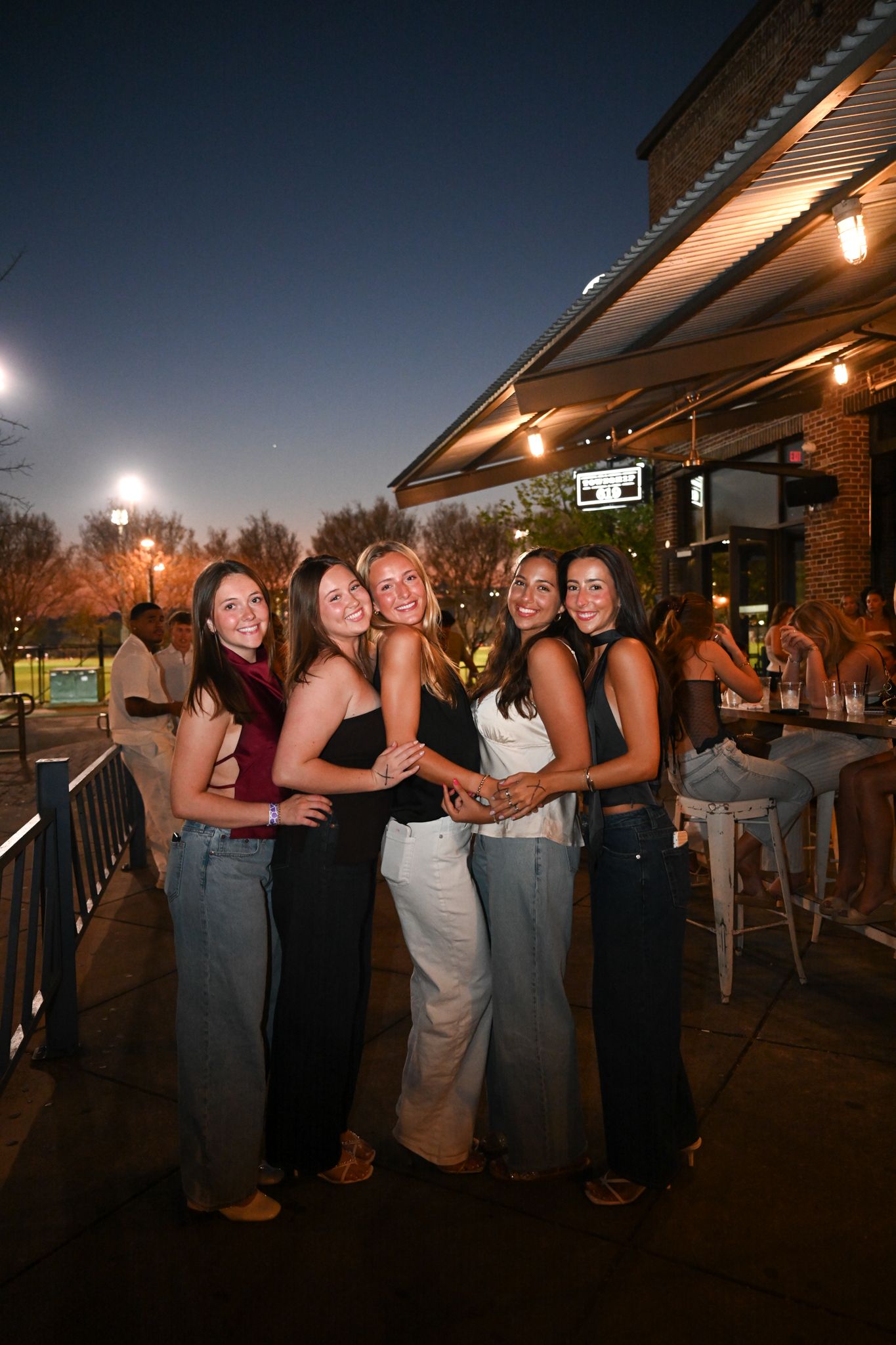 Five young women pose together smiling outside a bar patio at night with string lights glowing behind them.