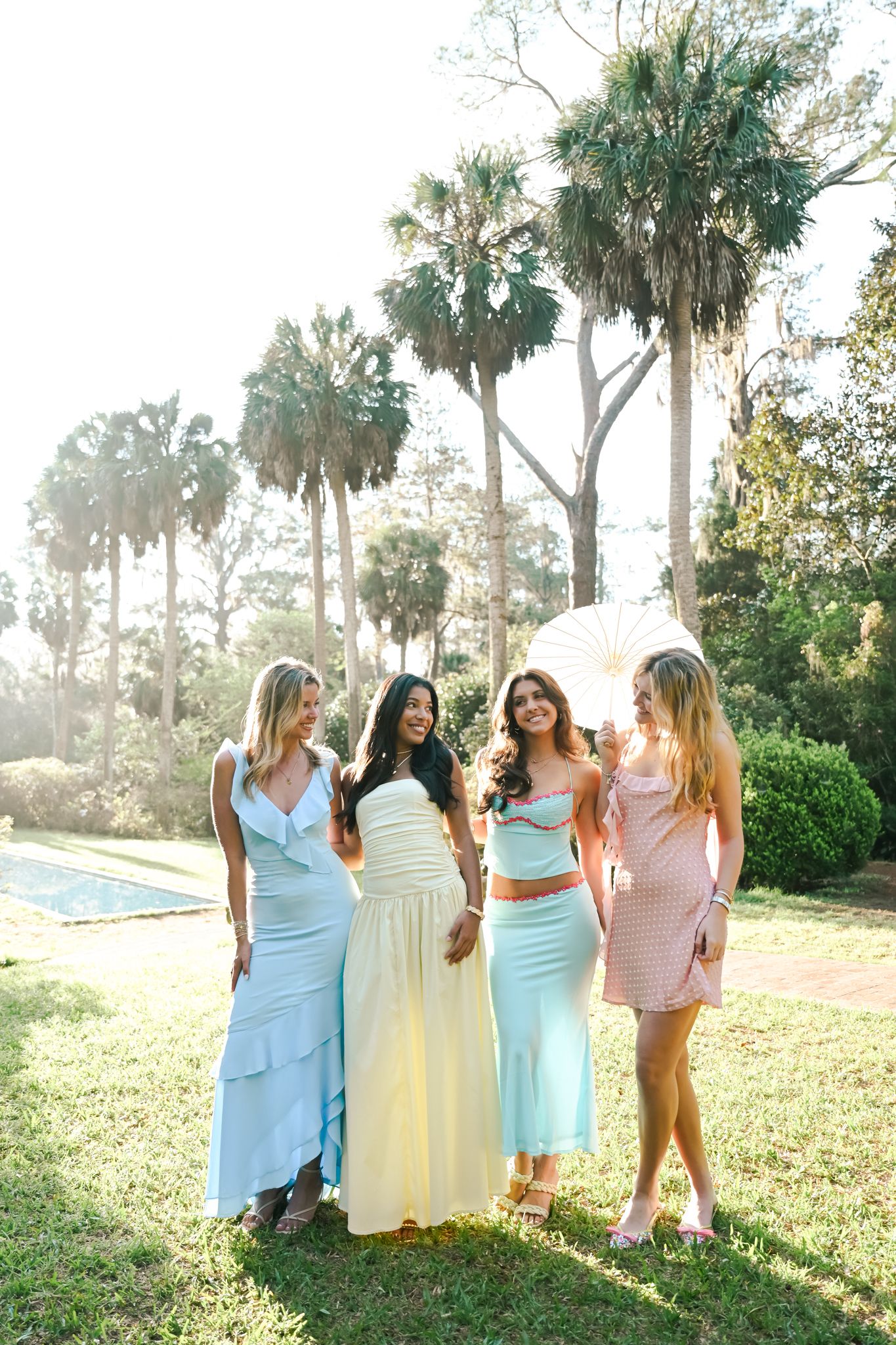 Four women in pastel summer dresses laughing together outdoors with palm trees and pool in background.