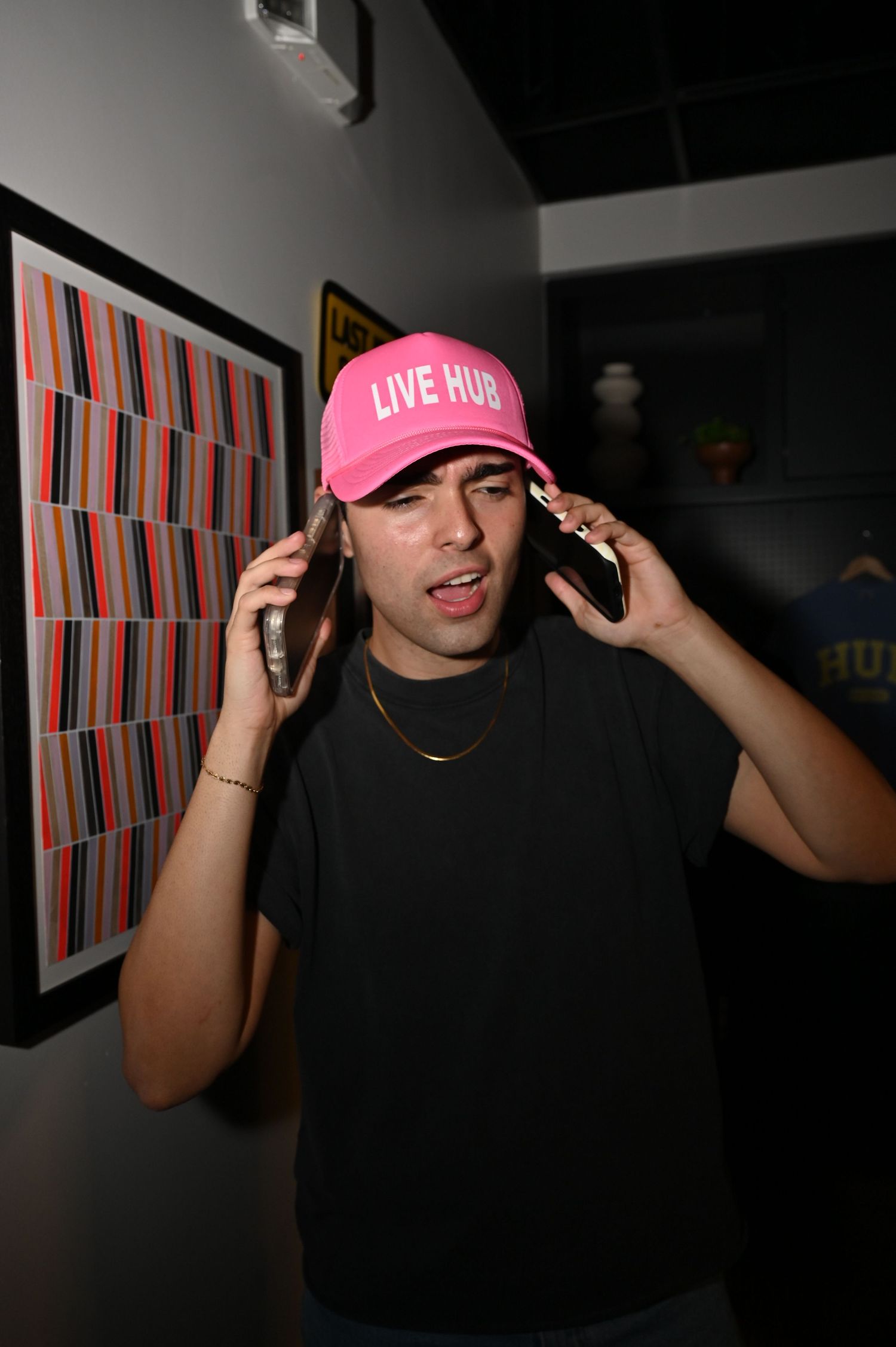 Man in pink Live Hub cap holds two phones to his ears near colorful striped artwork on wall.