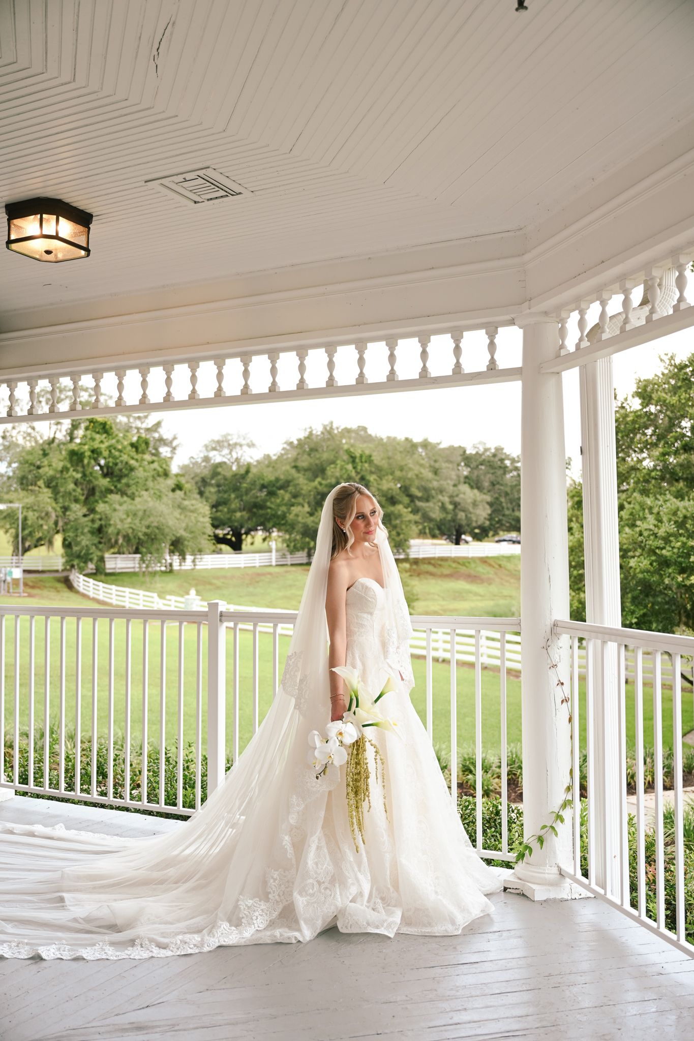 Bride in strapless lace ball gown with long veil holding floral bouquet on white southern plantation porch.