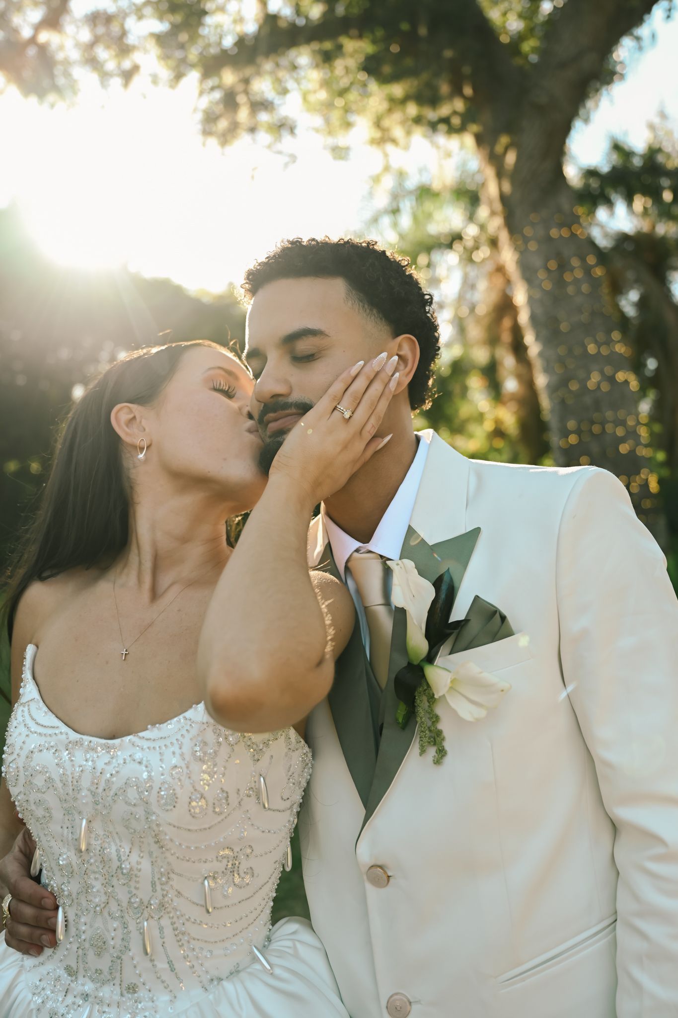 Bride in beaded gown kisses groom in ivory suit cheek during golden hour outdoor wedding ceremony.