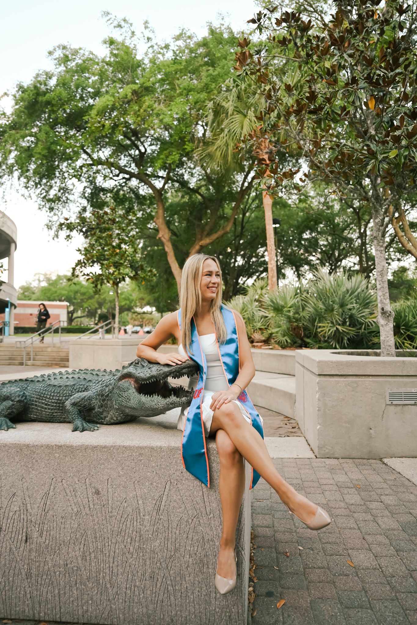 Smiling blonde woman in blue dress sitting on stone ledge beside alligator statue surrounded by lush green trees.