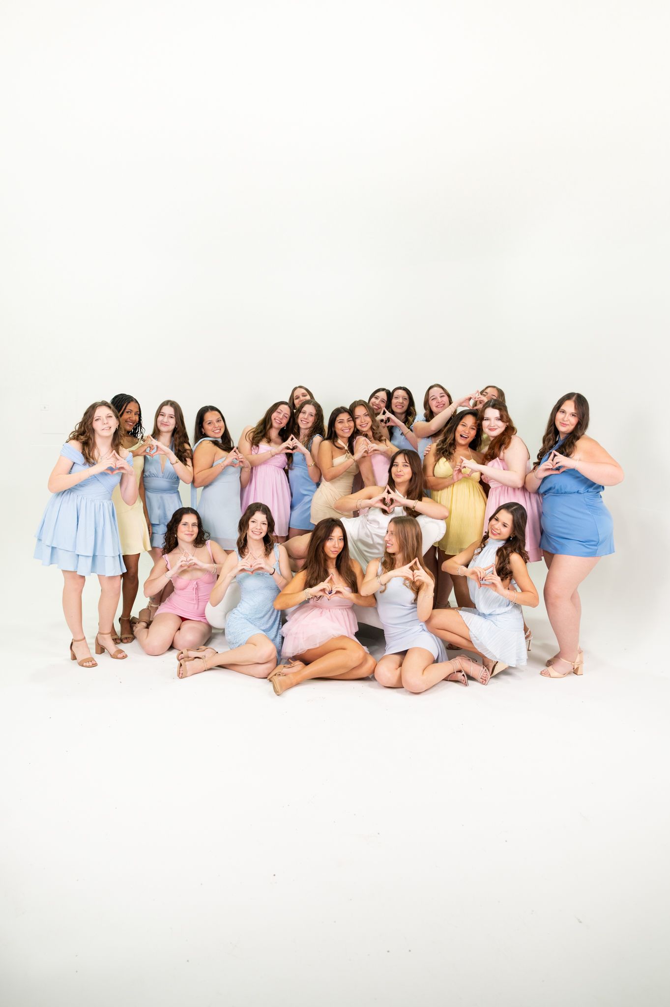Large group of young women in pastel dresses posing together, making heart shapes with their hands in a bright studio.