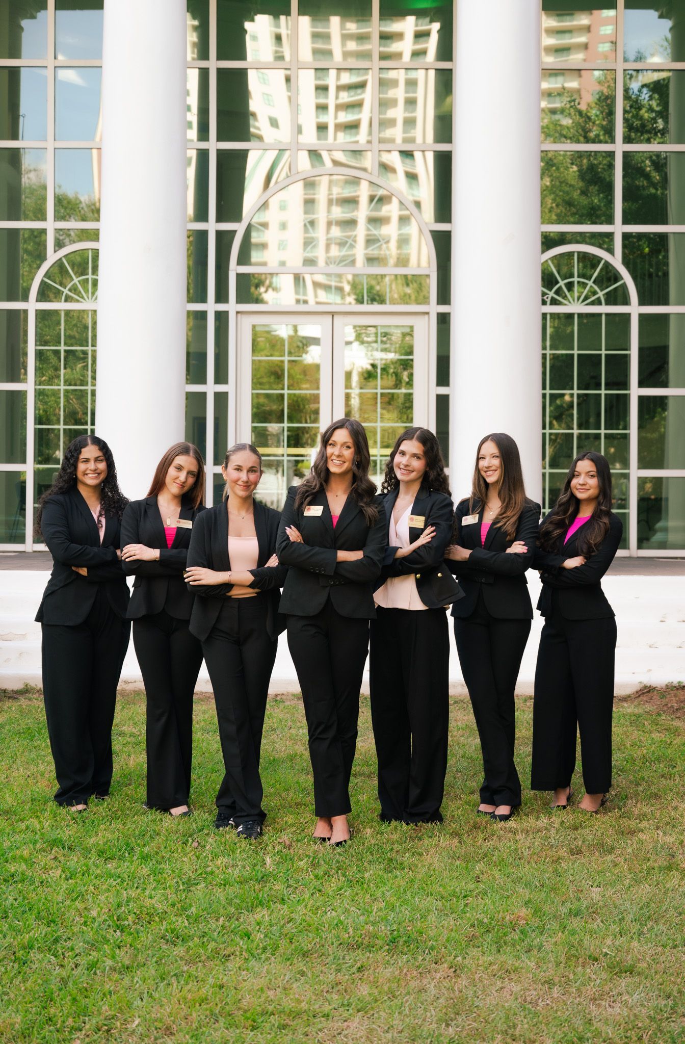 Seven women in black suits pose confidently with arms crossed in front of a elegant building with large windows.