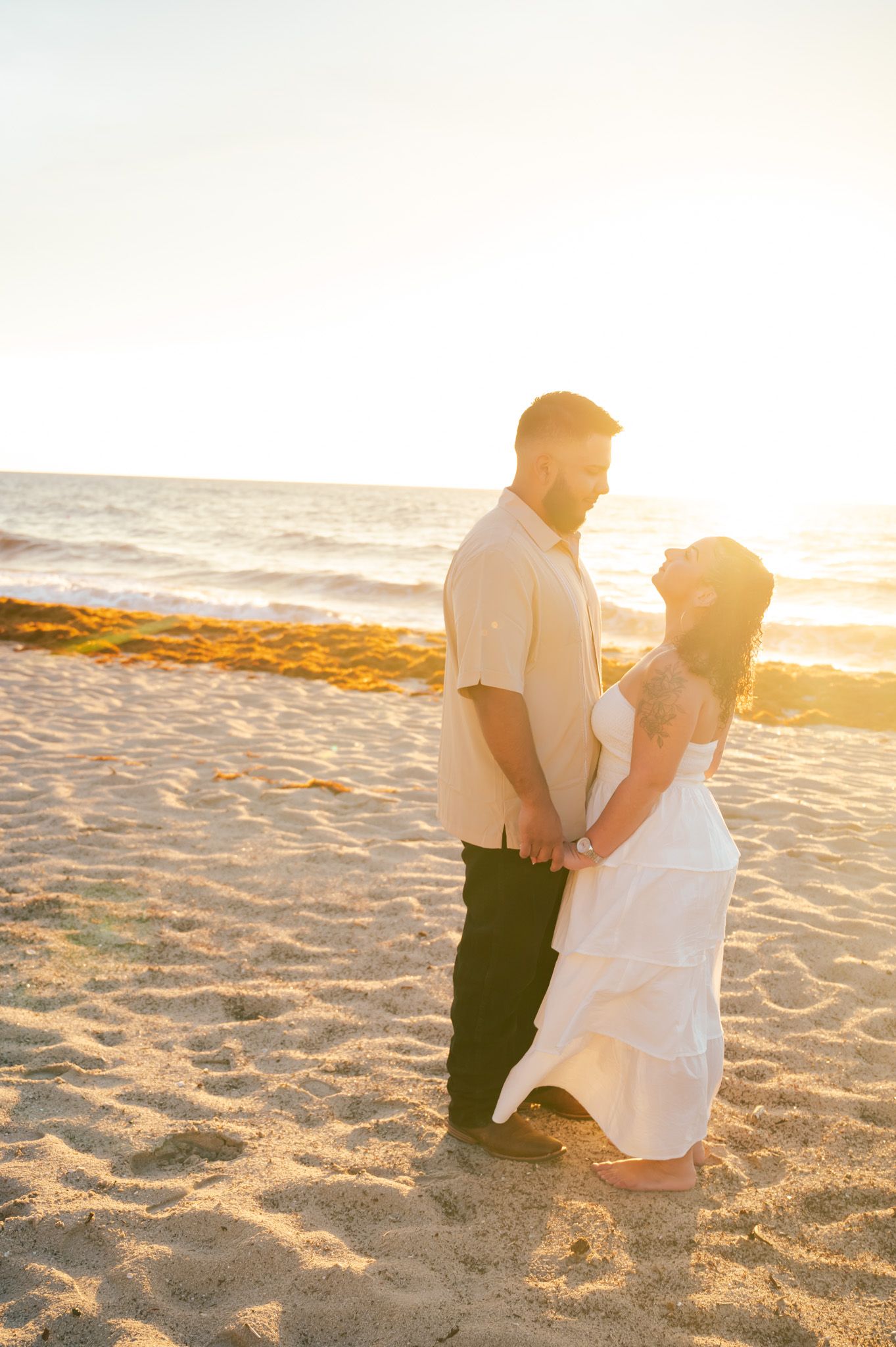 Couple holding hands on sandy beach at golden sunset, woman in white dress, man in beige shirt and black pants.