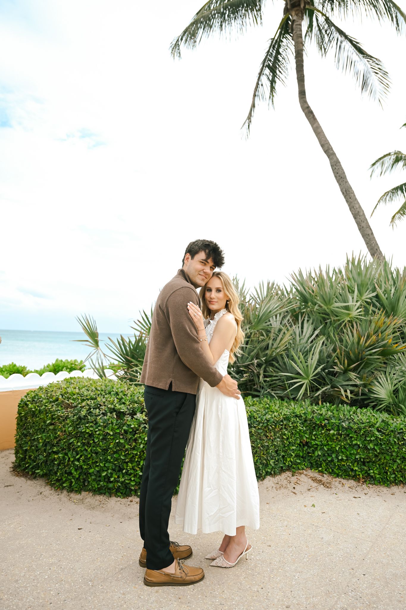 Couple embracing near tropical beach with palm tree and lush greenery in background.