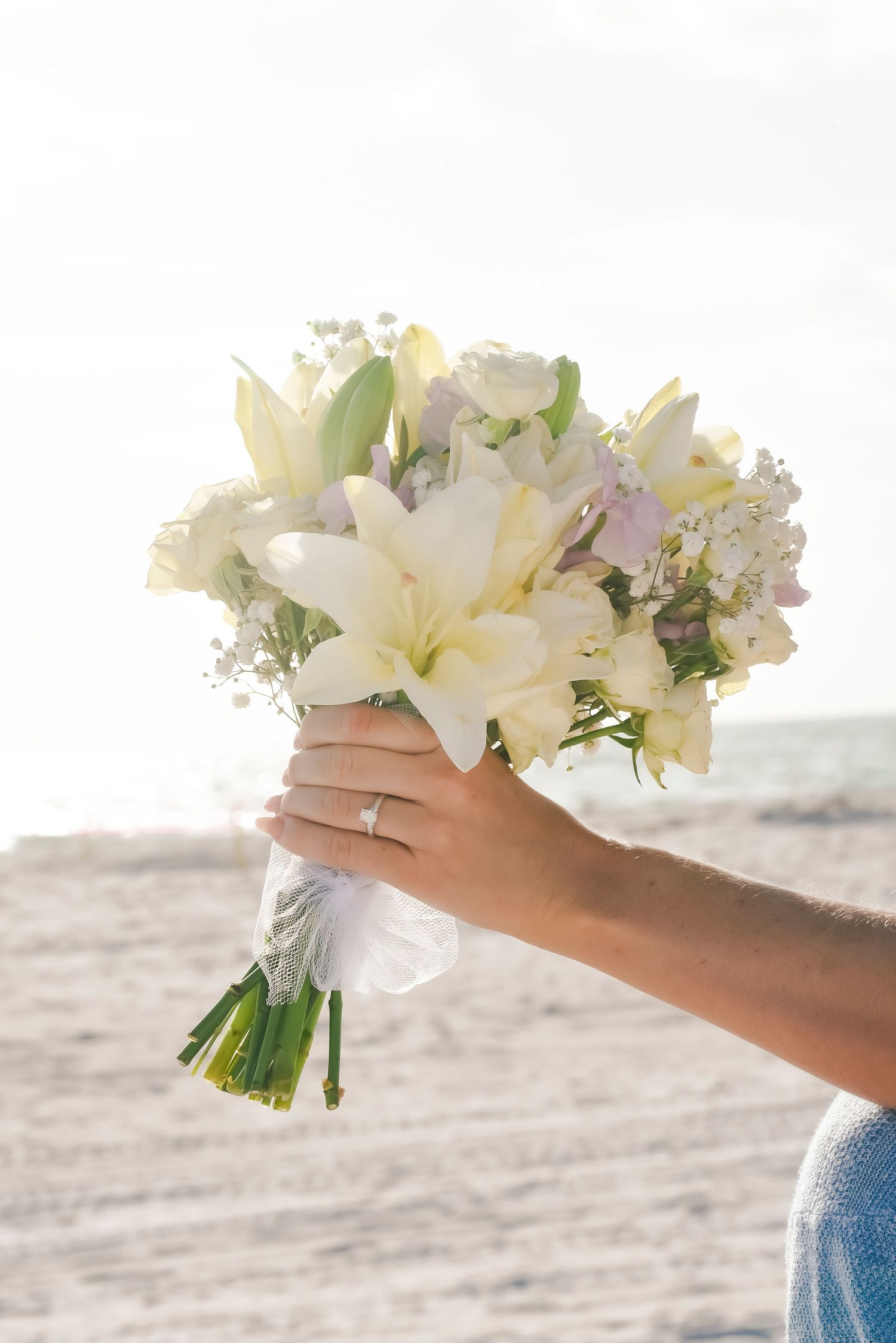 A bride holds a white lily and rose wedding bouquet on a sandy beach, wearing a diamond engagement ring.
