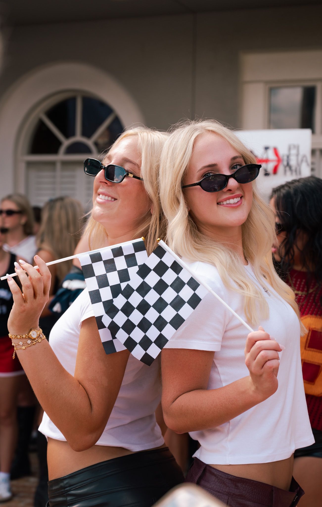 Two blonde women in white tees and sunglasses smile while holding checkered racing flags outdoors.