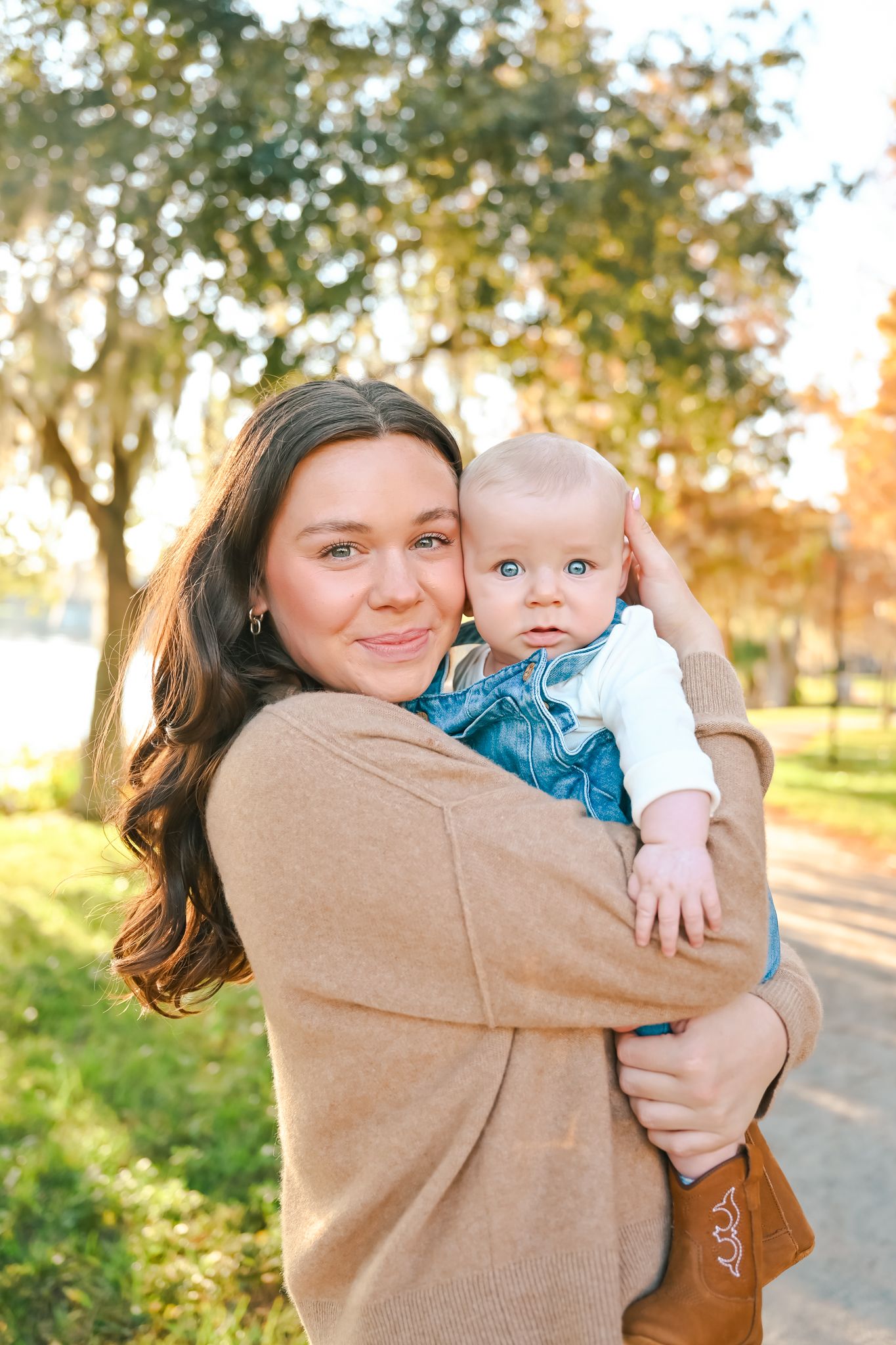 A person in a tan sweater holds and cuddles a baby wearing blue outdoors.