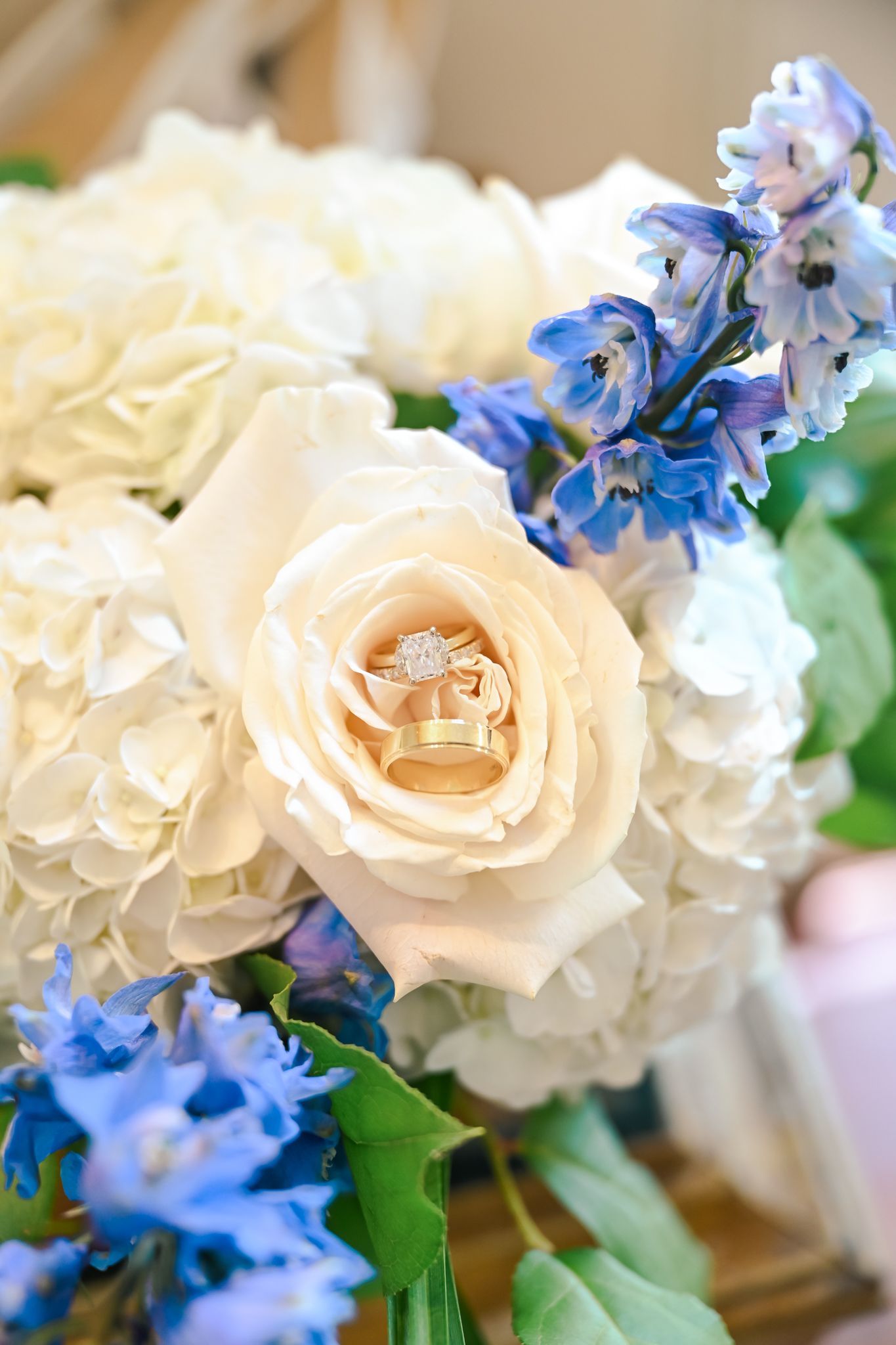 Wedding rings nestled in a cream rose bouquet with white hydrangeas and blue delphinium flowers.