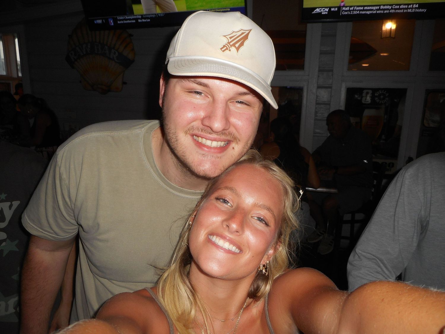 Two smiling young adults pose together indoors, woman with blonde hair in front, man wearing white cap behind her.