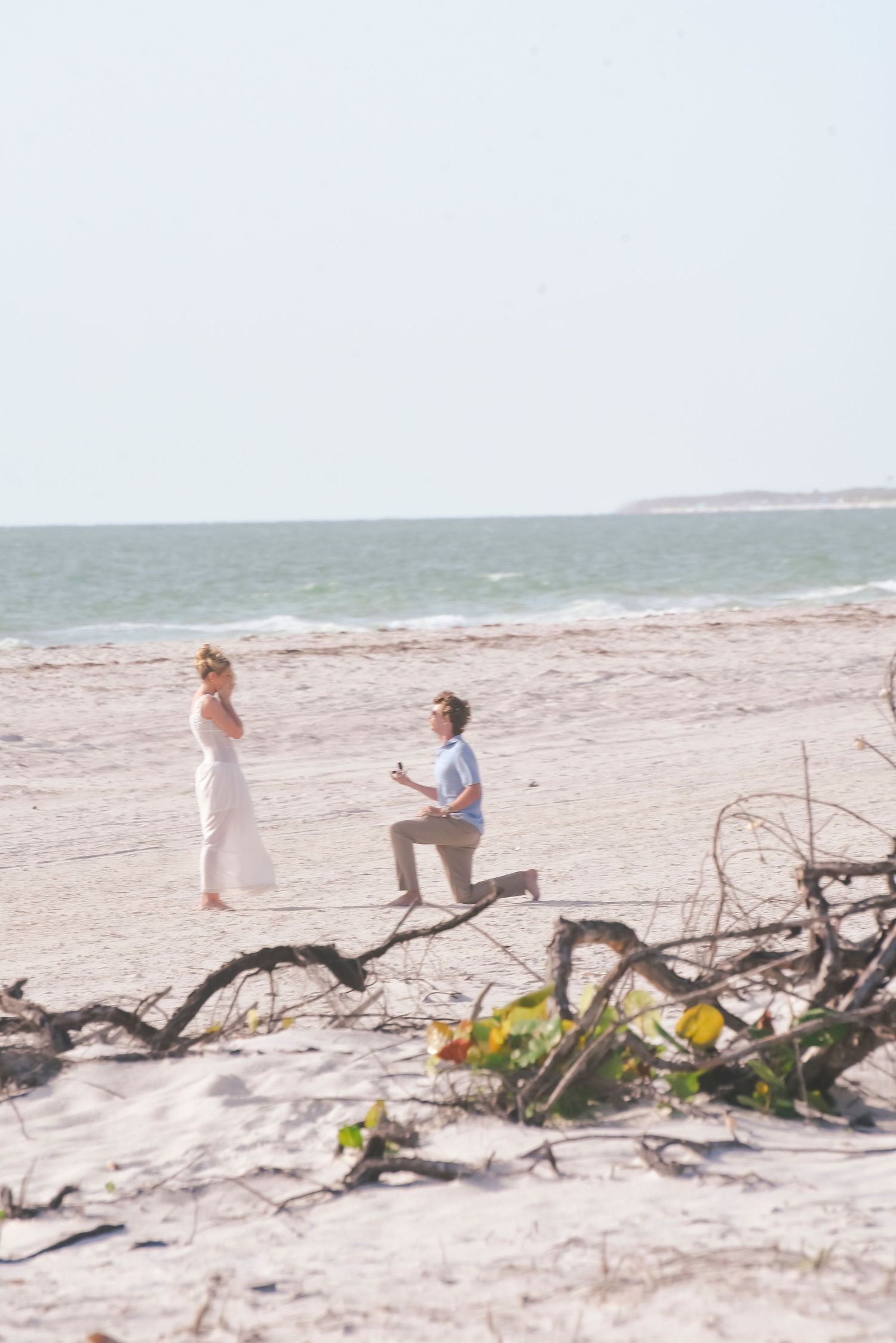 Man proposing on one knee to woman in white dress on sandy beach with ocean waves in background.