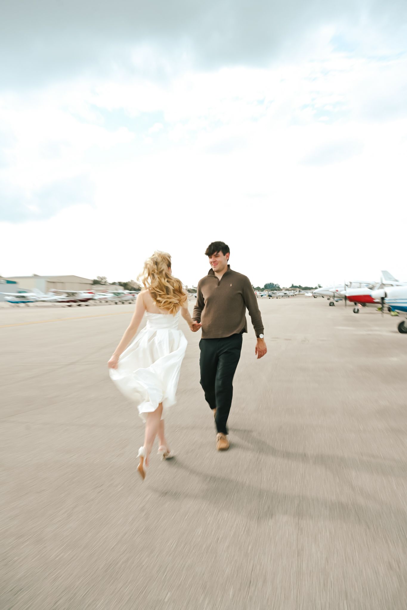 Couple holding hands walking on airport tarmac, woman in white dress, man in dark pants, planes visible in background.
