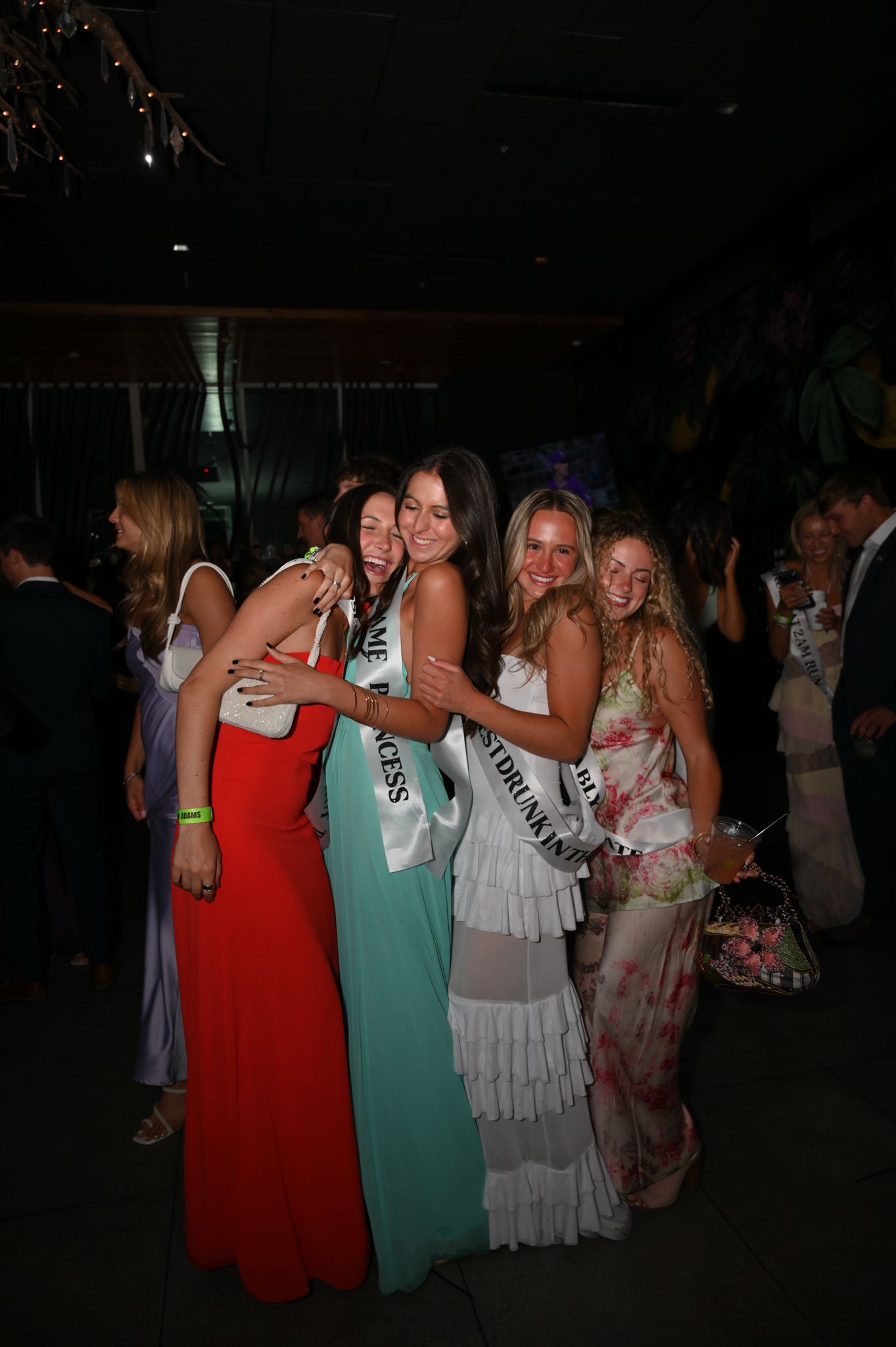 Four women in formal gowns, two wearing pageant sashes, pose together smiling at an elegant evening event.