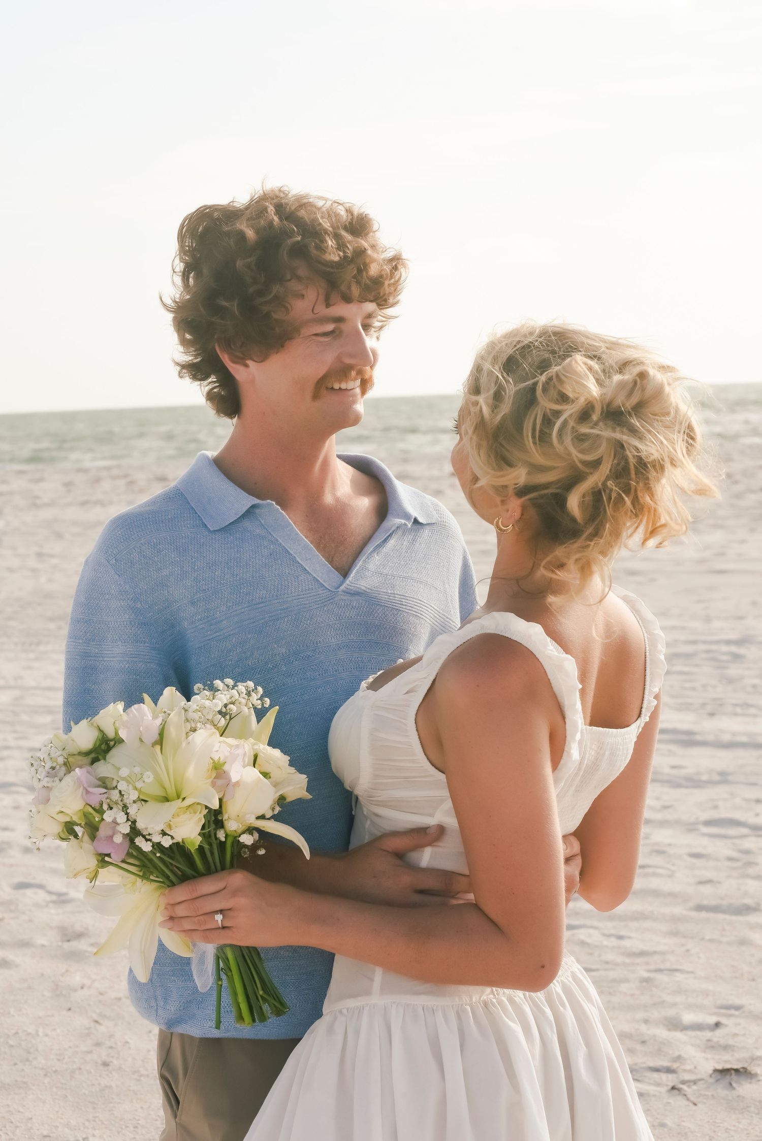 Couple shares intimate moment on beach with white floral bouquet at sunset.