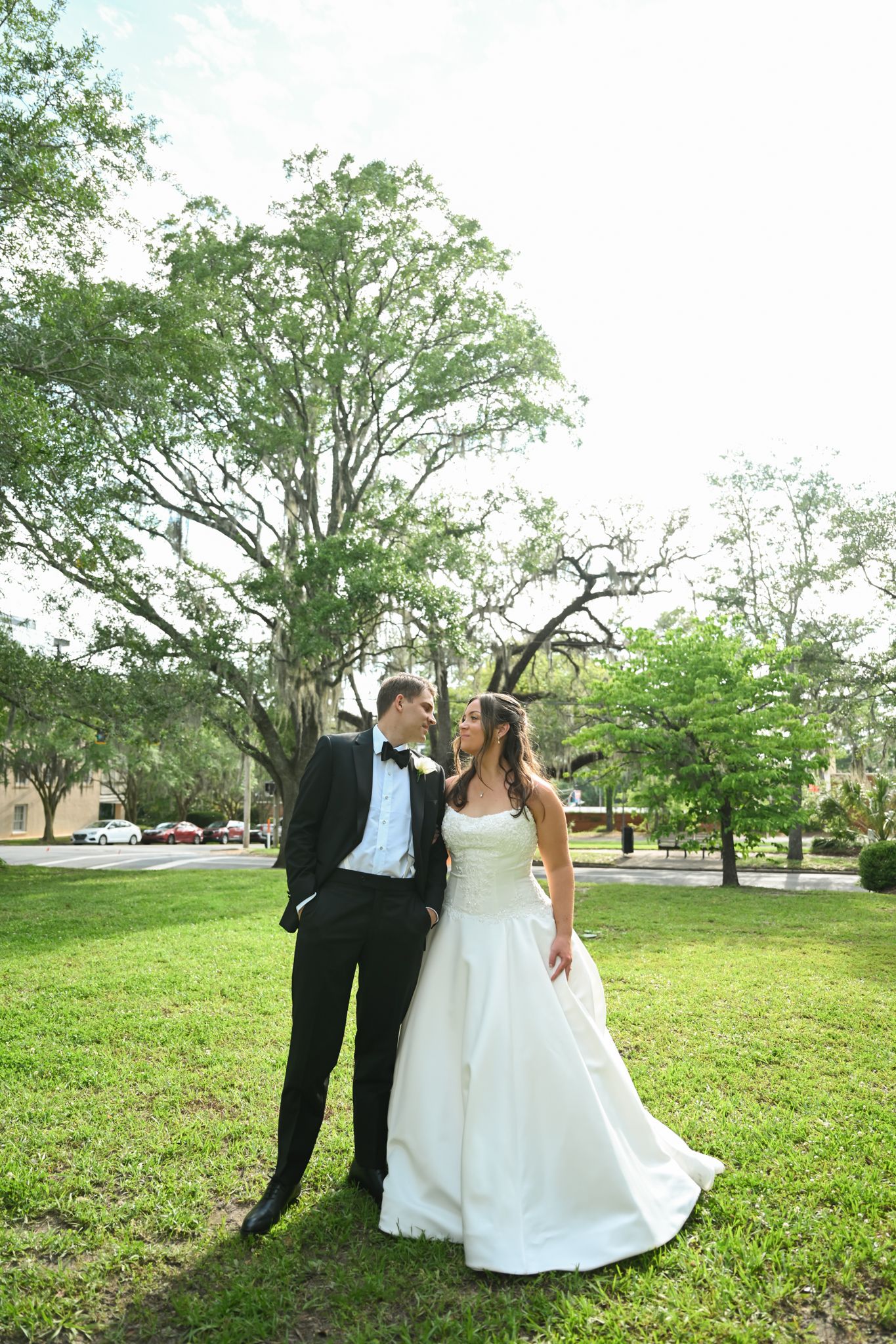 Bride and groom share a romantic moment outdoors on lush green grass beneath a large oak tree on their wedding day.