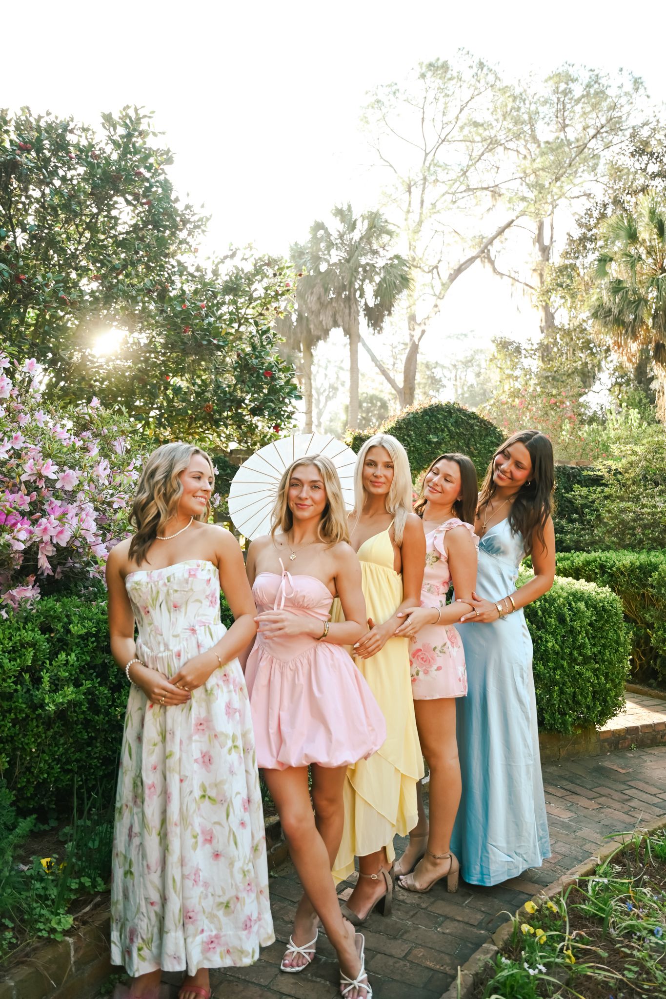 Five women in pastel dresses pose in a sunlit garden, one holding a white parasol.
