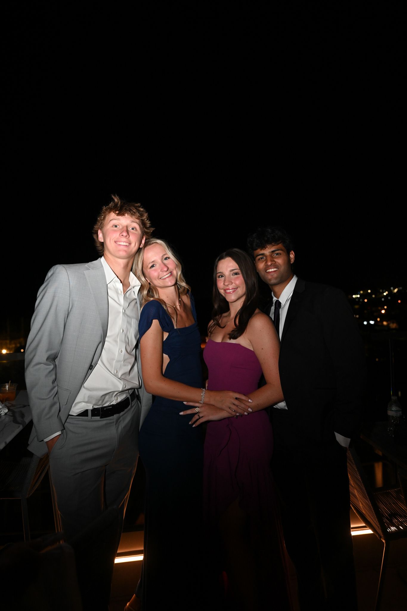 Two couples dressed in formal attire smile together at an outdoor evening event.