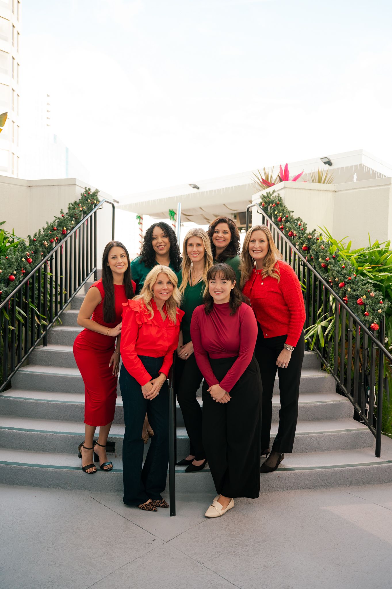 Seven women in red and green outfits pose on outdoor stairs decorated with holiday garland.