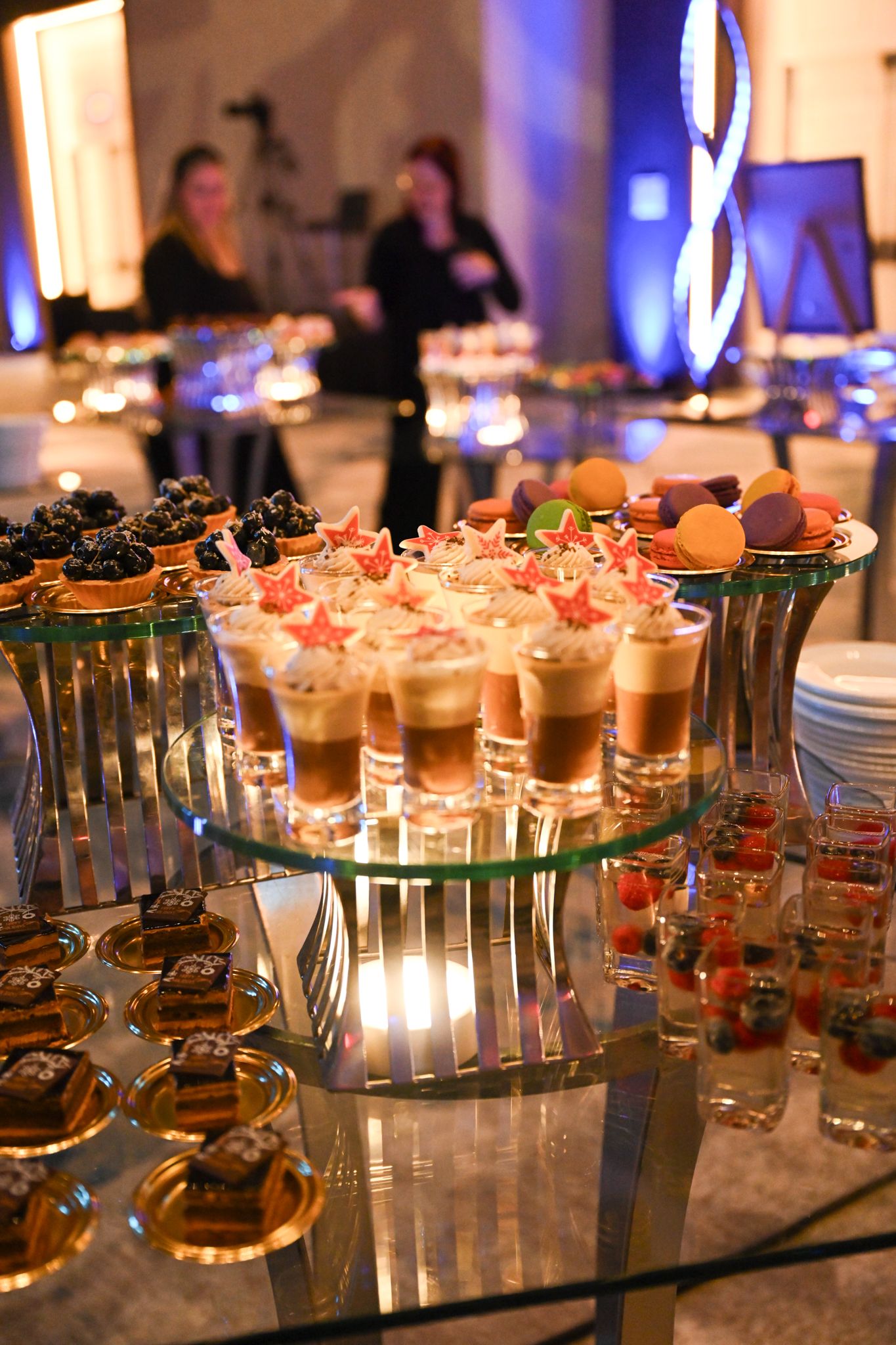 Elegant dessert buffet display with layered mousse shots, macarons, and berry tarts on glass tiers at a catered event.