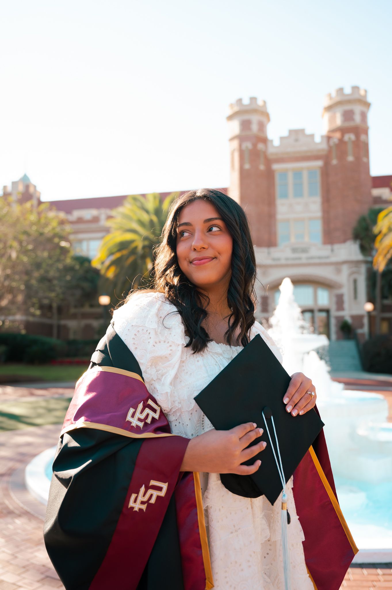 Graduate in maroon and white regalia poses joyfully in front of campus fountain.