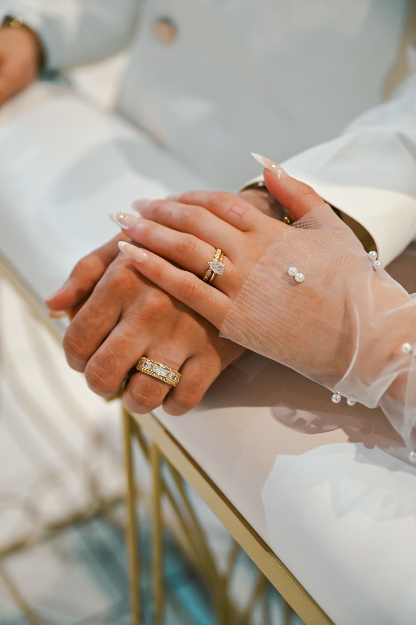 Close-up of bride and groom hands adorned with gold wedding rings and pearl bracelet in elegant white attire.