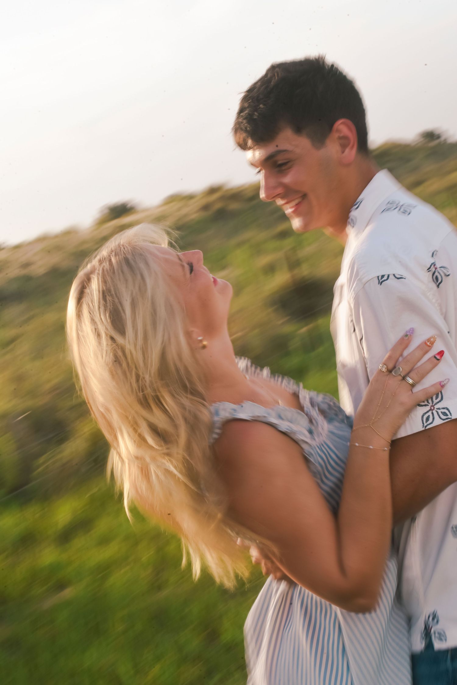 A smiling young couple embraces outdoors in a sunny green field, sharing a joyful romantic moment.