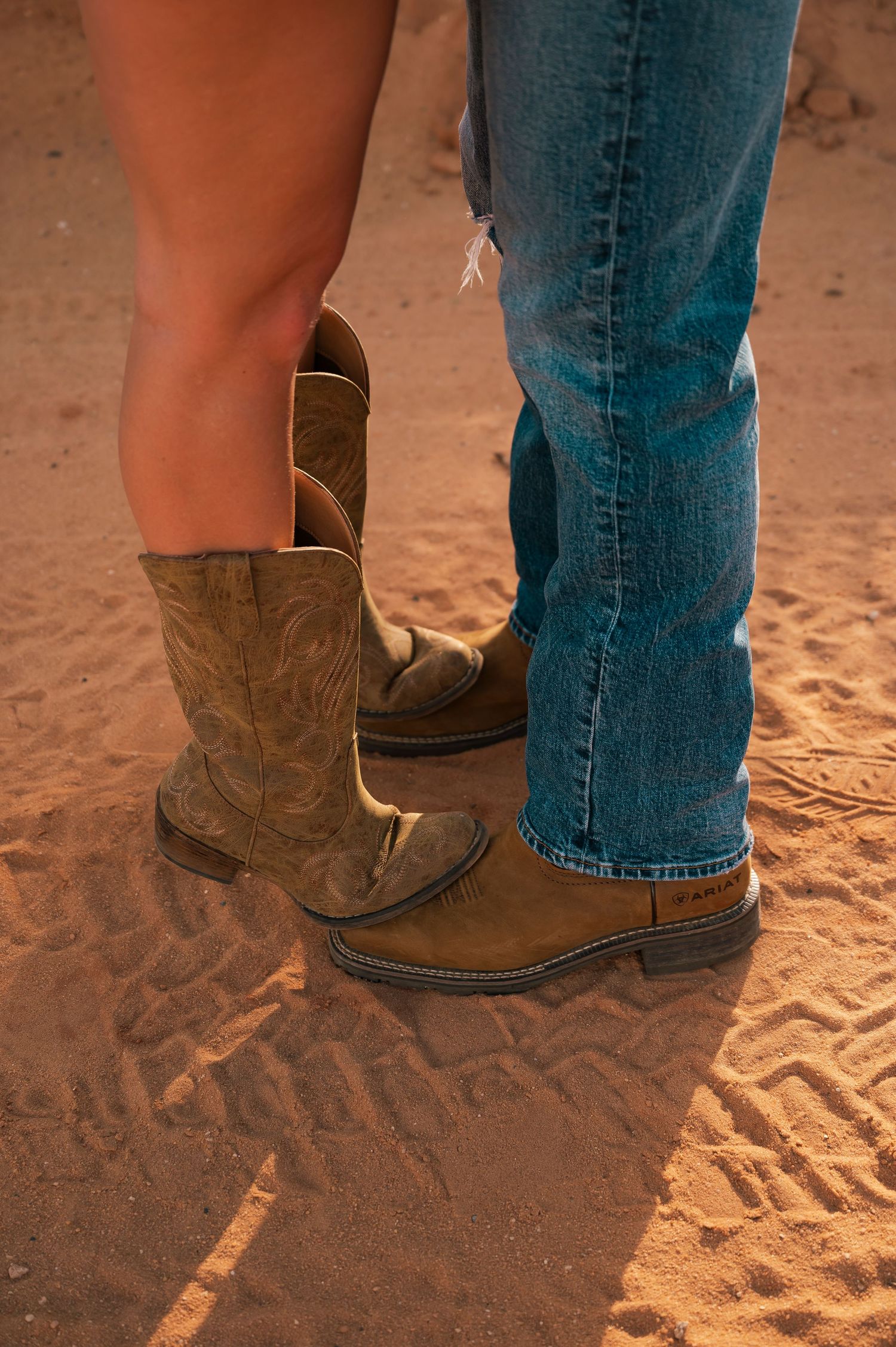 Two people wearing matching tan cowboy boots standing on red sandy ground, one in jeans and one with bare legs.