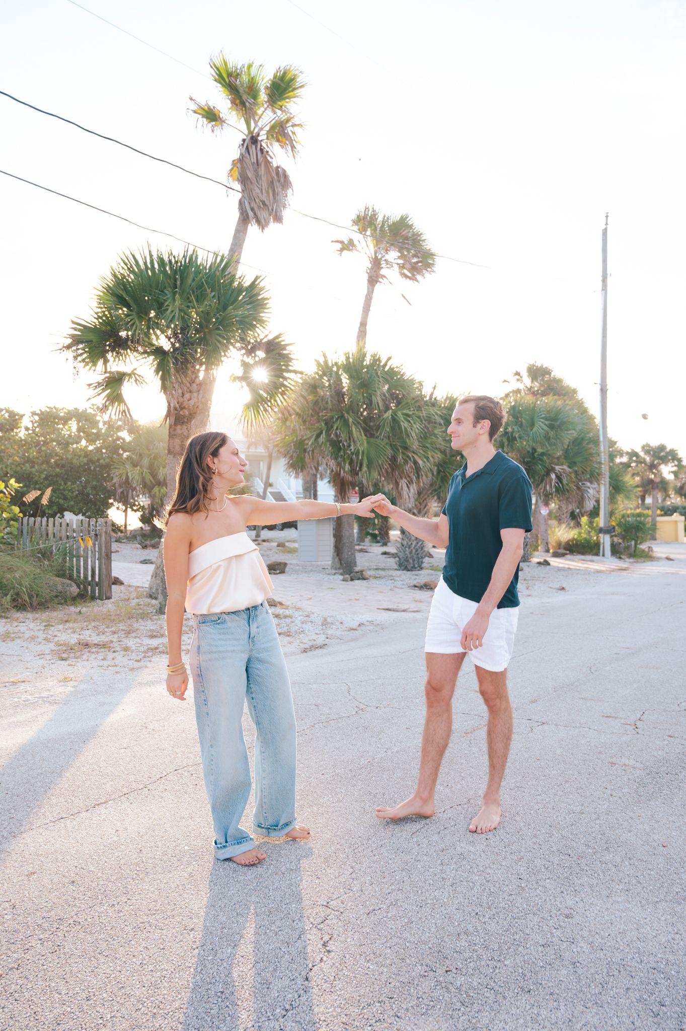 Couple dancing on a sunny street lined with palm trees, woman in white top and jeans, man in navy shirt and white shorts.