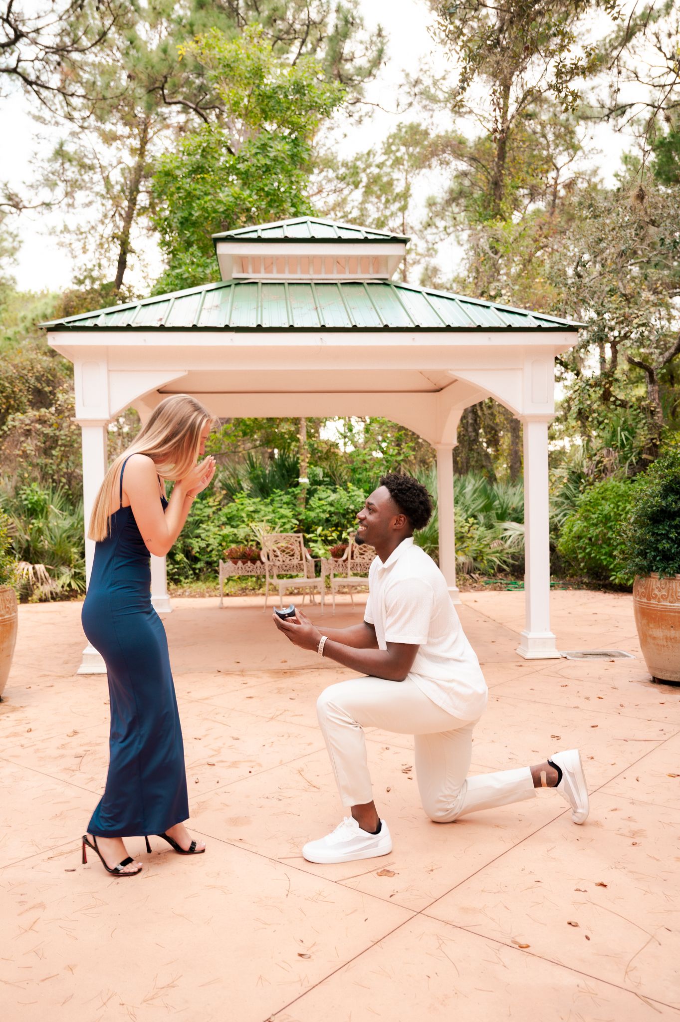Man proposes on one knee to woman in blue dress near white gazebo in lush garden setting.